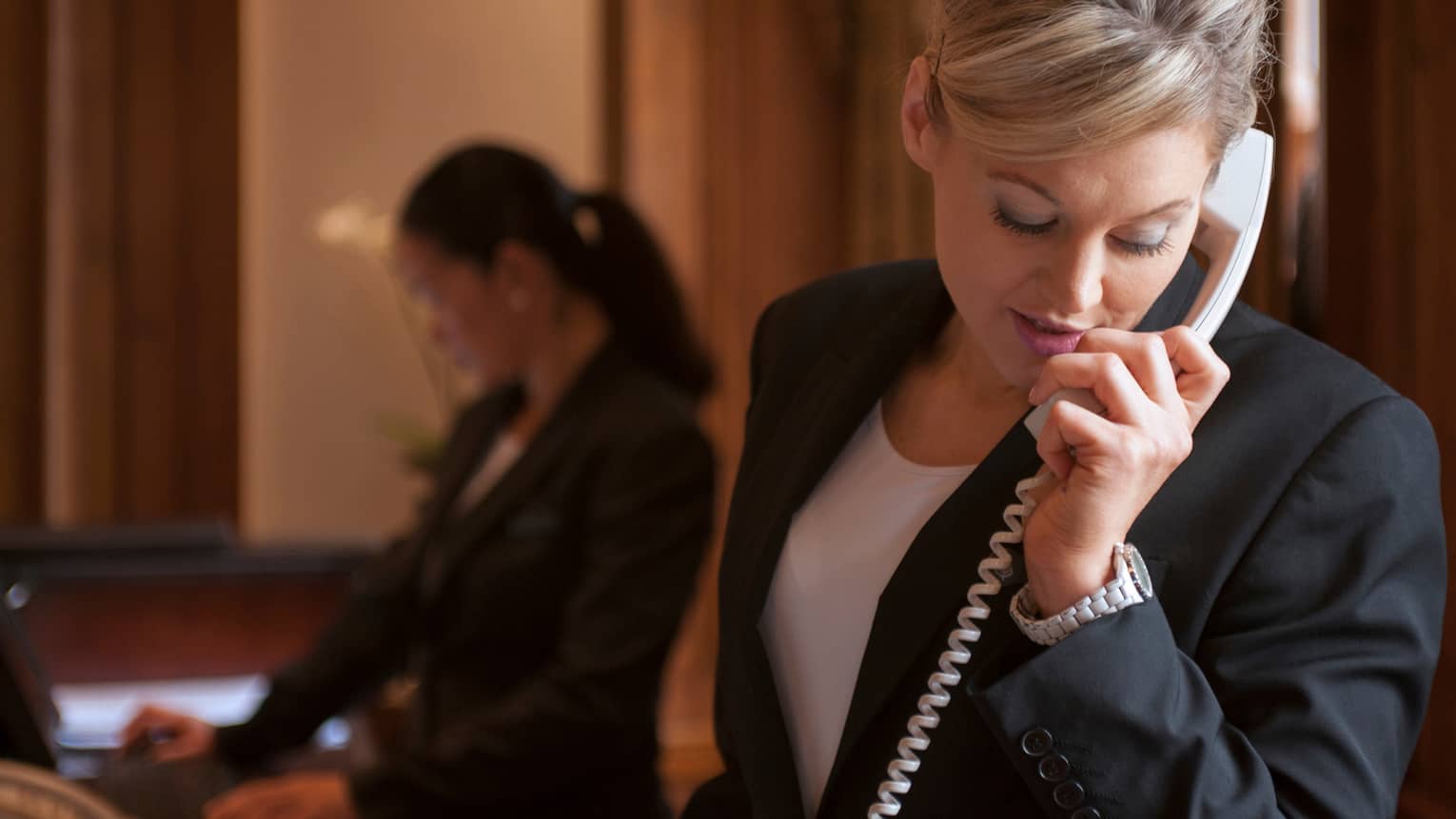 Close-up of woman in dark suit jacket holding telephone, cord, silhouette of hotel reception staff in background