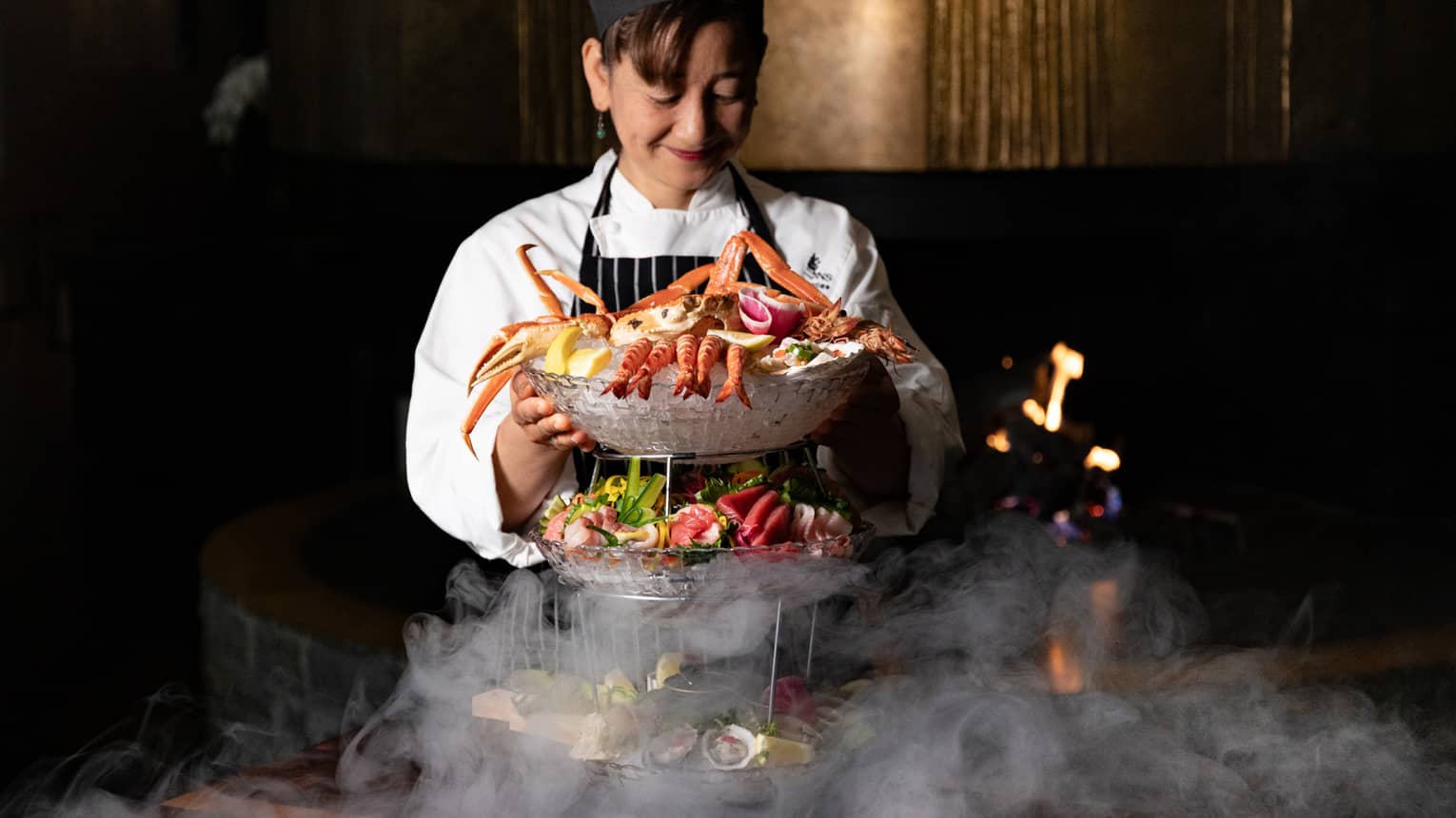 Female chef wearing black chef's hat, white chef's coat and black-and-white pinstripe apron smiles as she sets up a three-layered seafood tower featuring crab legs and assortment of seafood items
