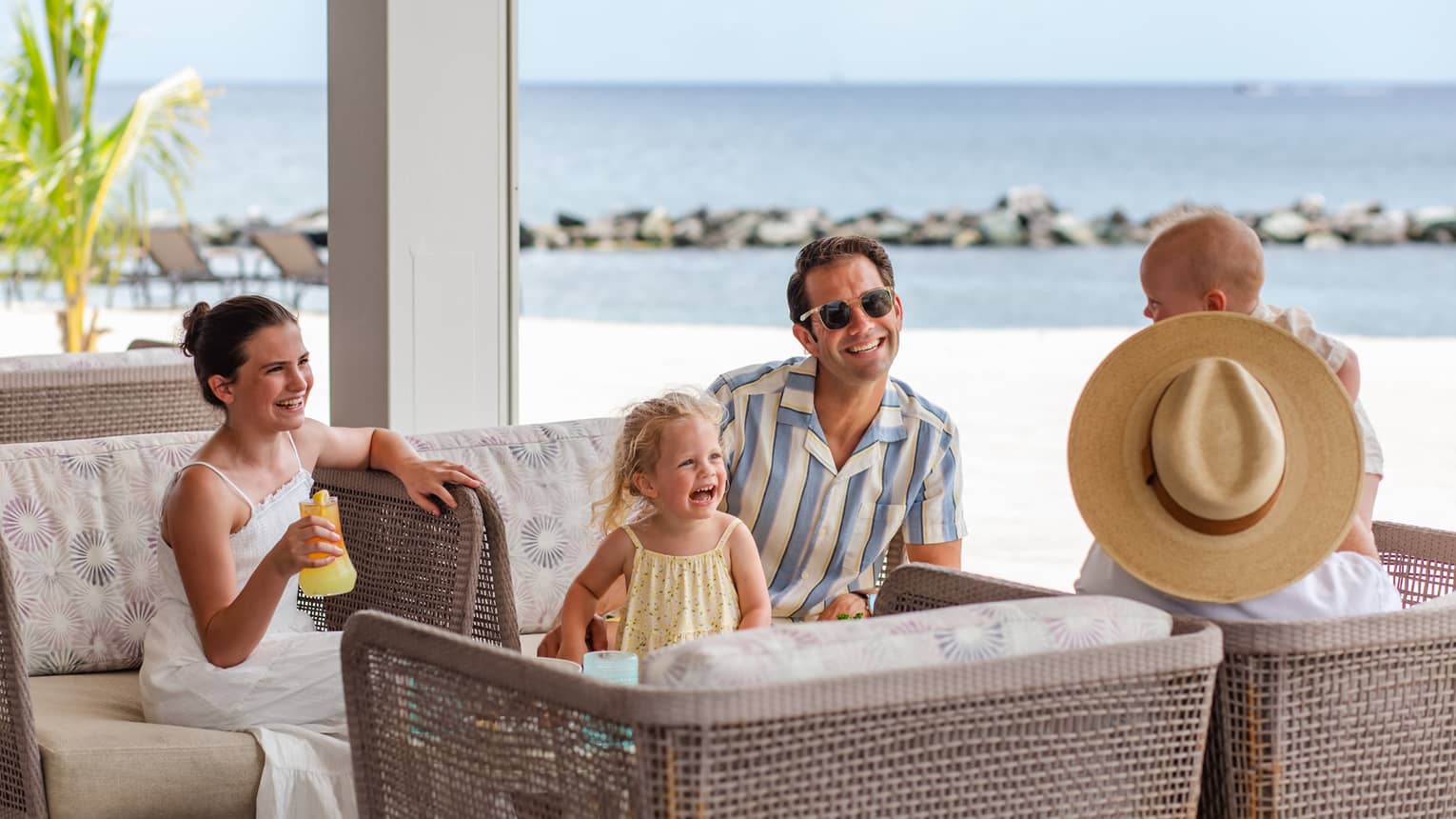 Three adults and two young children sit together facing each other on four rattan arm chairs with a beach in the background