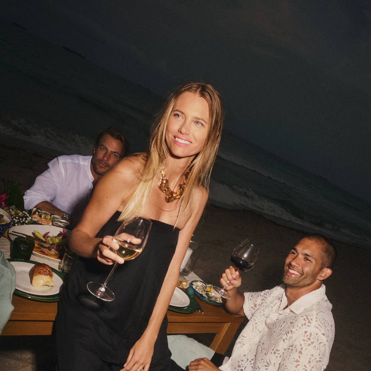 Guests sit around a table set on the beach, smiling and drinking red wine
