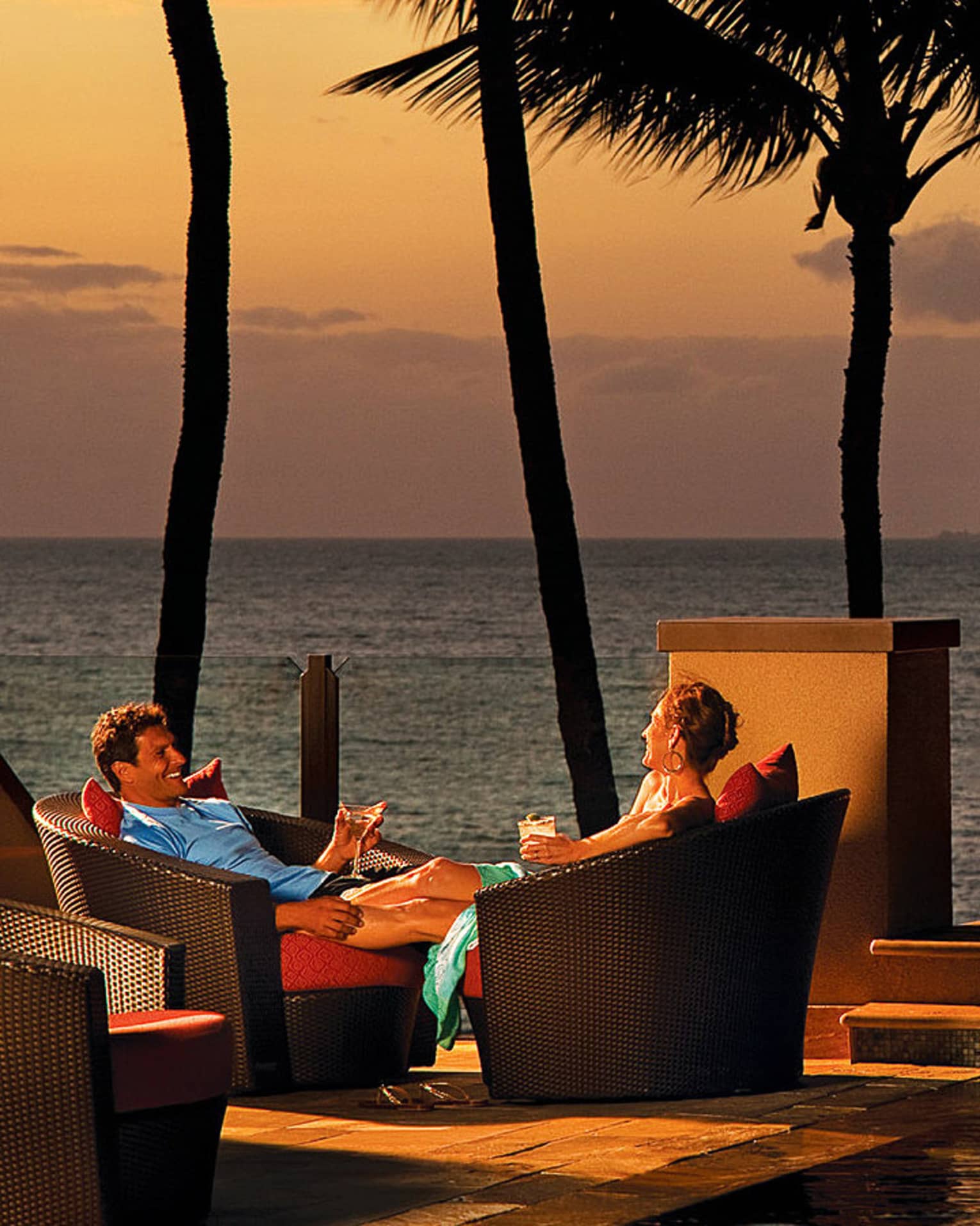 Man and woman sit in wicker patio chairs with cocktails at sunset, next to infinity pool and fire pit