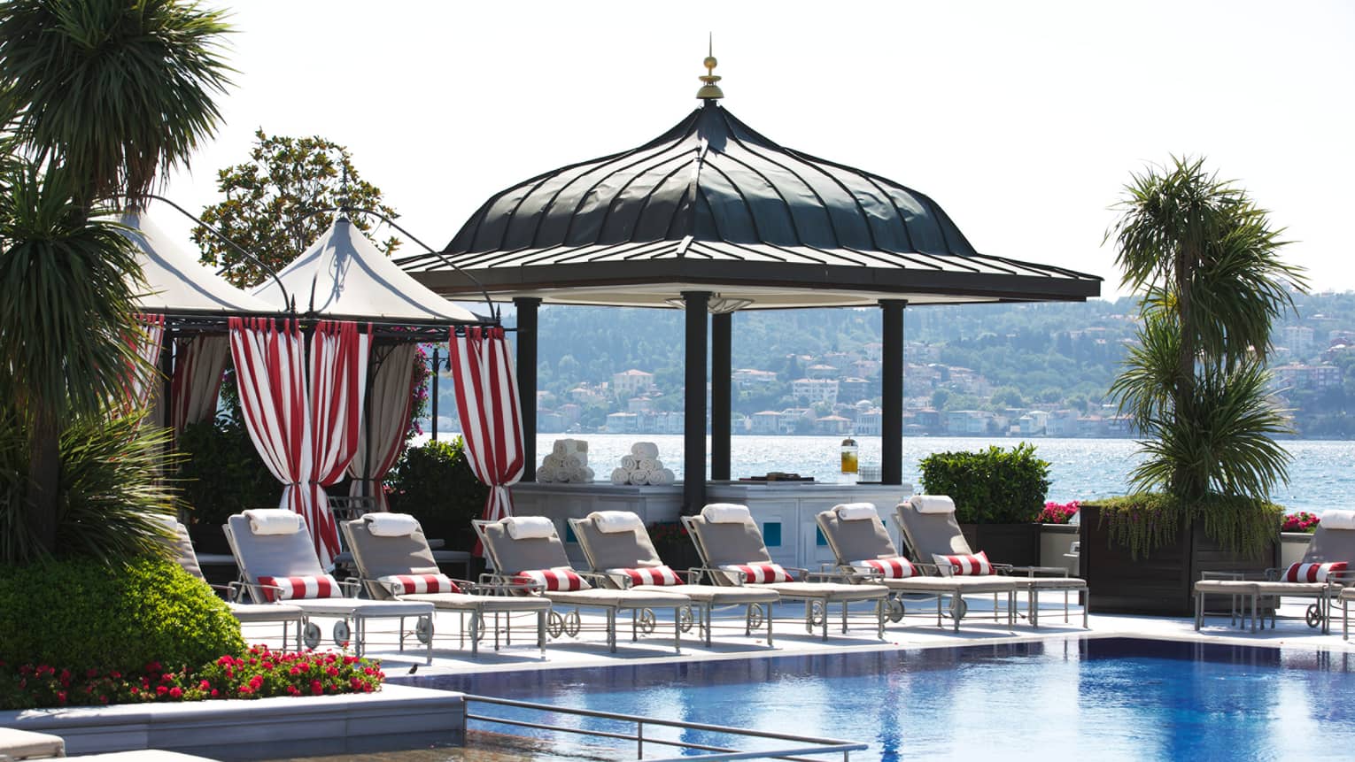 Outdoor pool lined with lounge chairs, red-and-white-striped towels rolled up, pavilion, sea in background