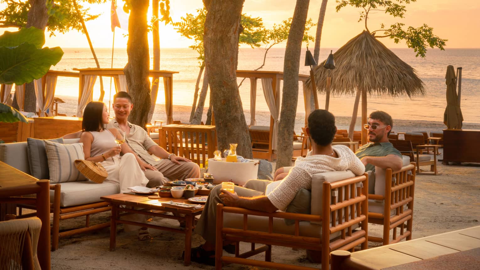 Four people sit around a table in rattan lounge chairs overlooking the beach at sunset