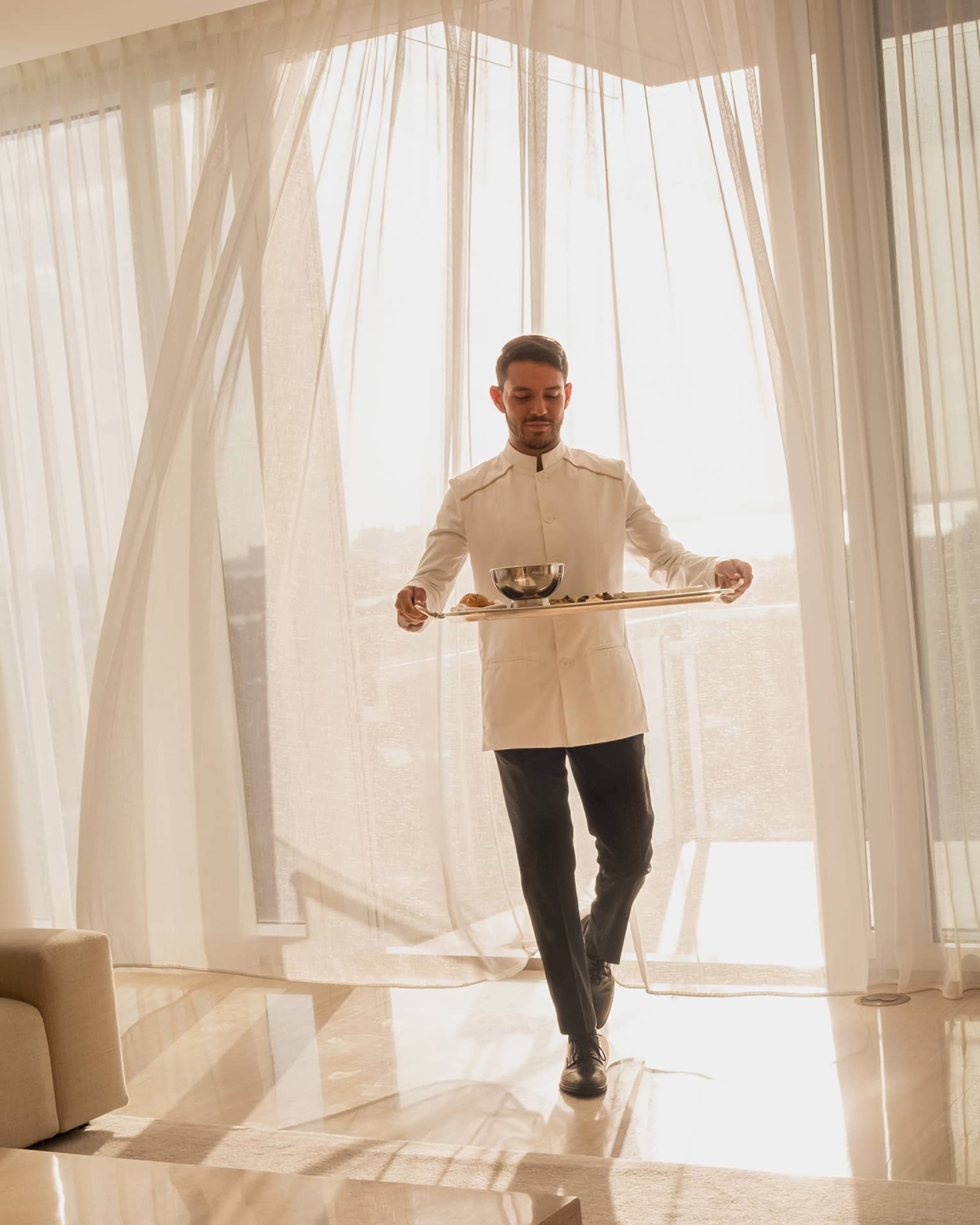 Hotel staff member carrying a tray with food for in-room dining, standing in front of sheer curtains with sunlight streaming through