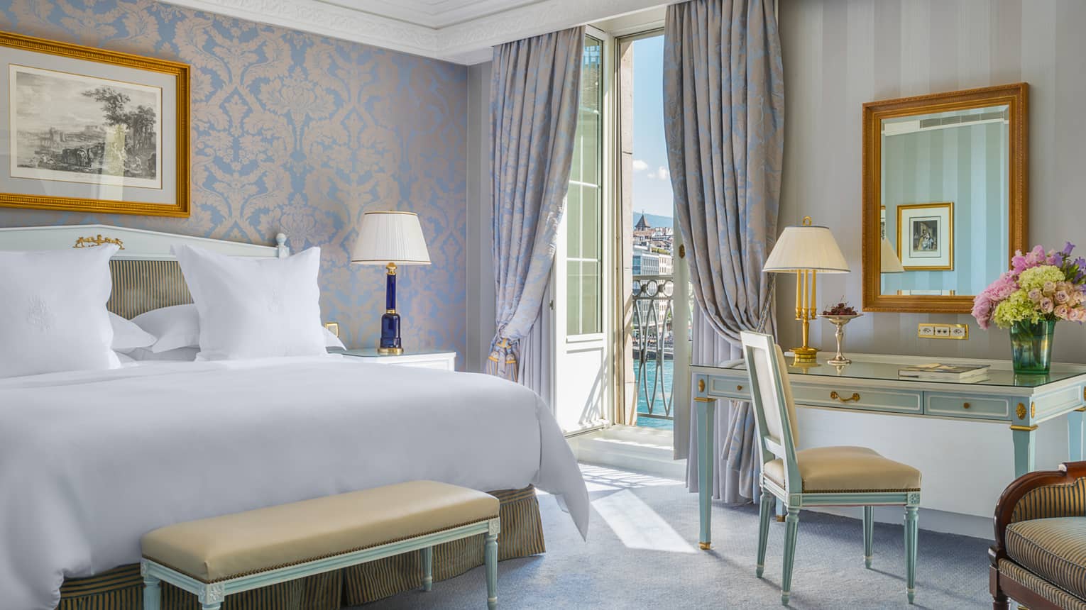 Elegant hotel room with a king bed, desk, seating area and a balcony overlooking water, featuring light blue and gold decor