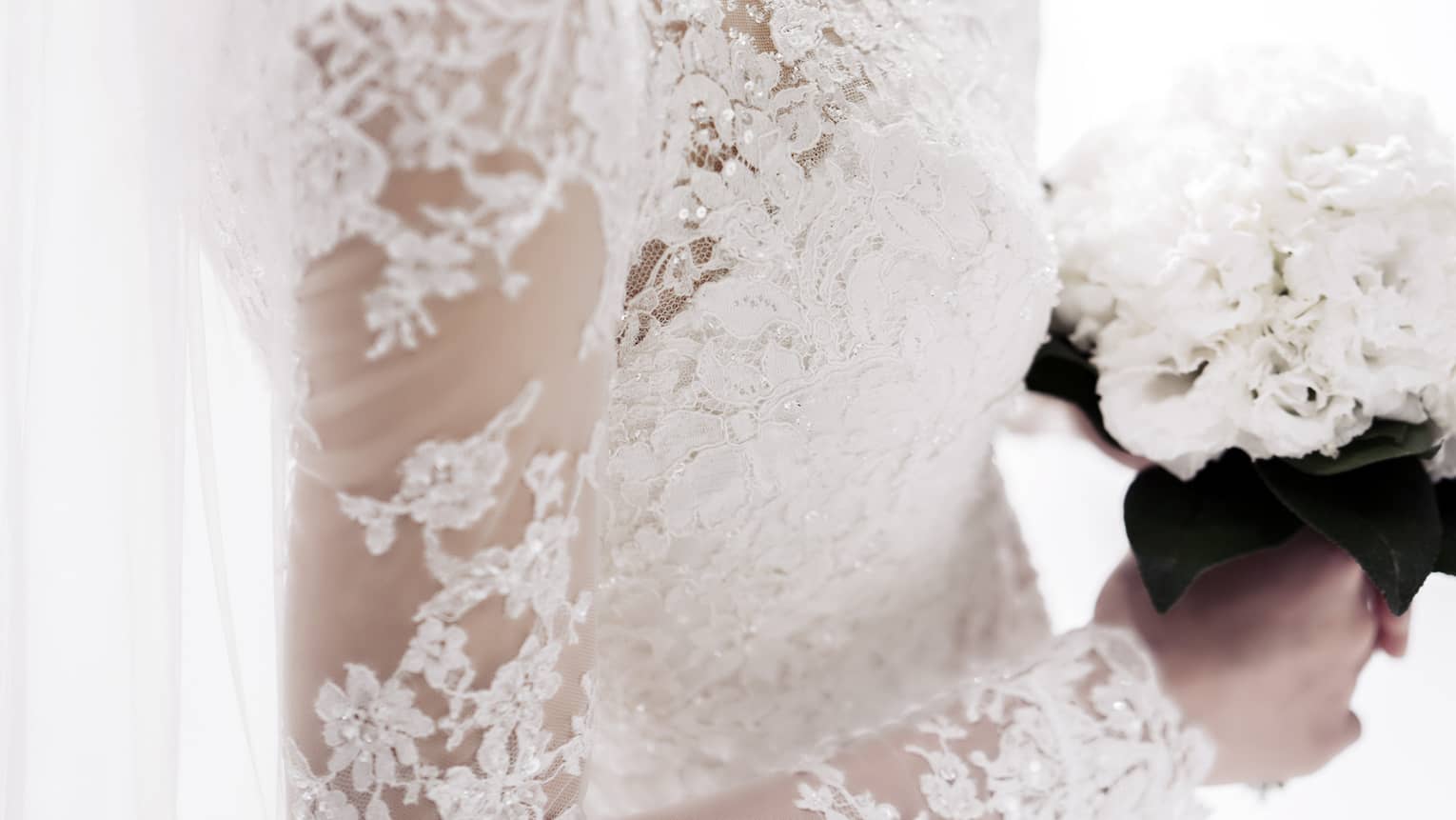 Over-the-shoulder view of a bride wearing a long-sleeved white lace wedding gown, a long organza veil and holding a bouquet of all-white flowers with dark leaves