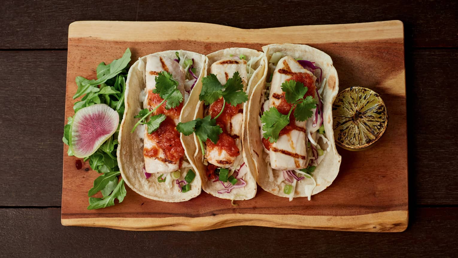 Aerial view of three large fish tacos, sauce on rustic wood platter