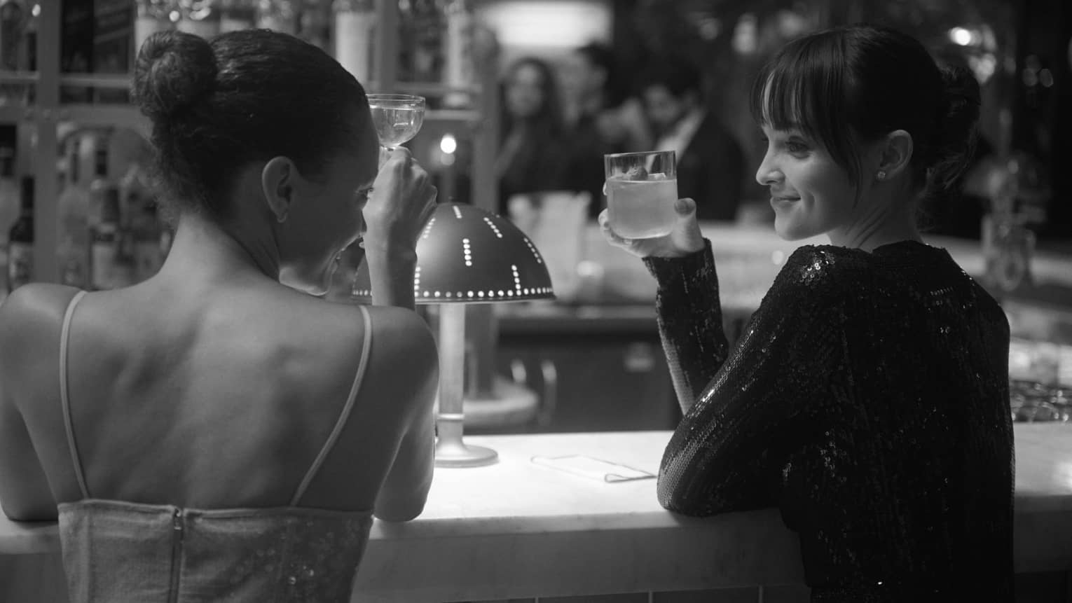 A black and white shot of two elegantly dressed guests at a bar, smiling and raising their cocktails.