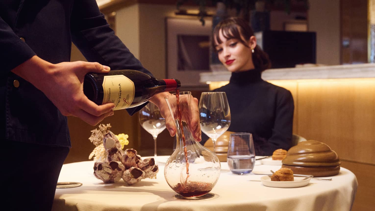 Suited server decants red wine at table with seated guest watching