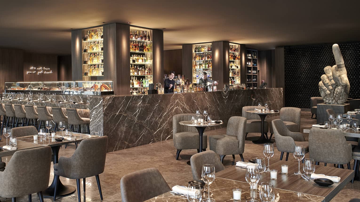 Bartenders behind shiny bar with columns lined with spirits and lounge club-style seating area with neutral decor