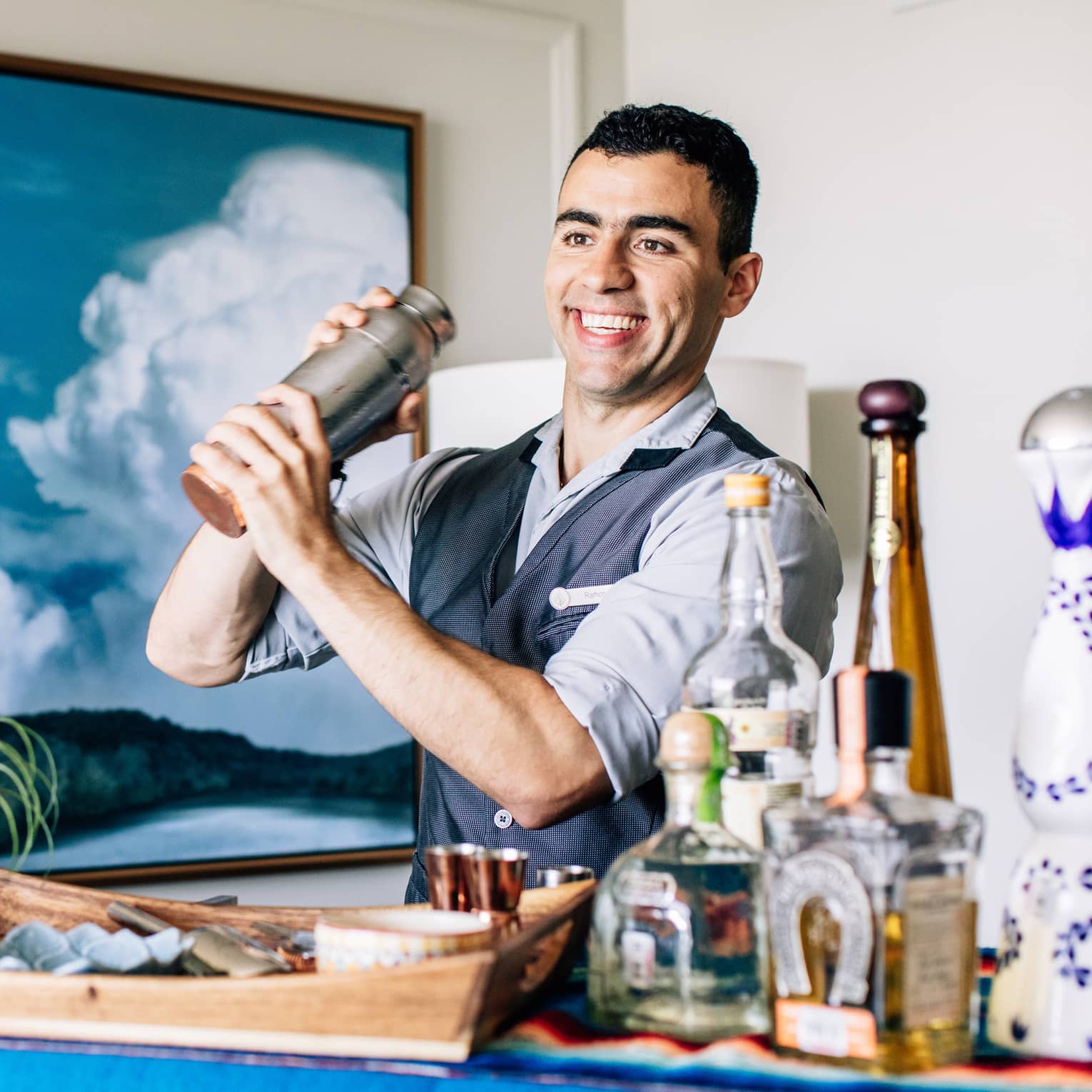 A bartender shaking a cocktail with liquor bottles on a cart in front of him.
