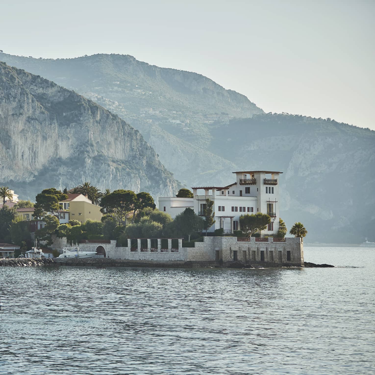 Cliffs and hills loom above a forested village along the water; a speed boat is anchored in front of a stone breakwater.