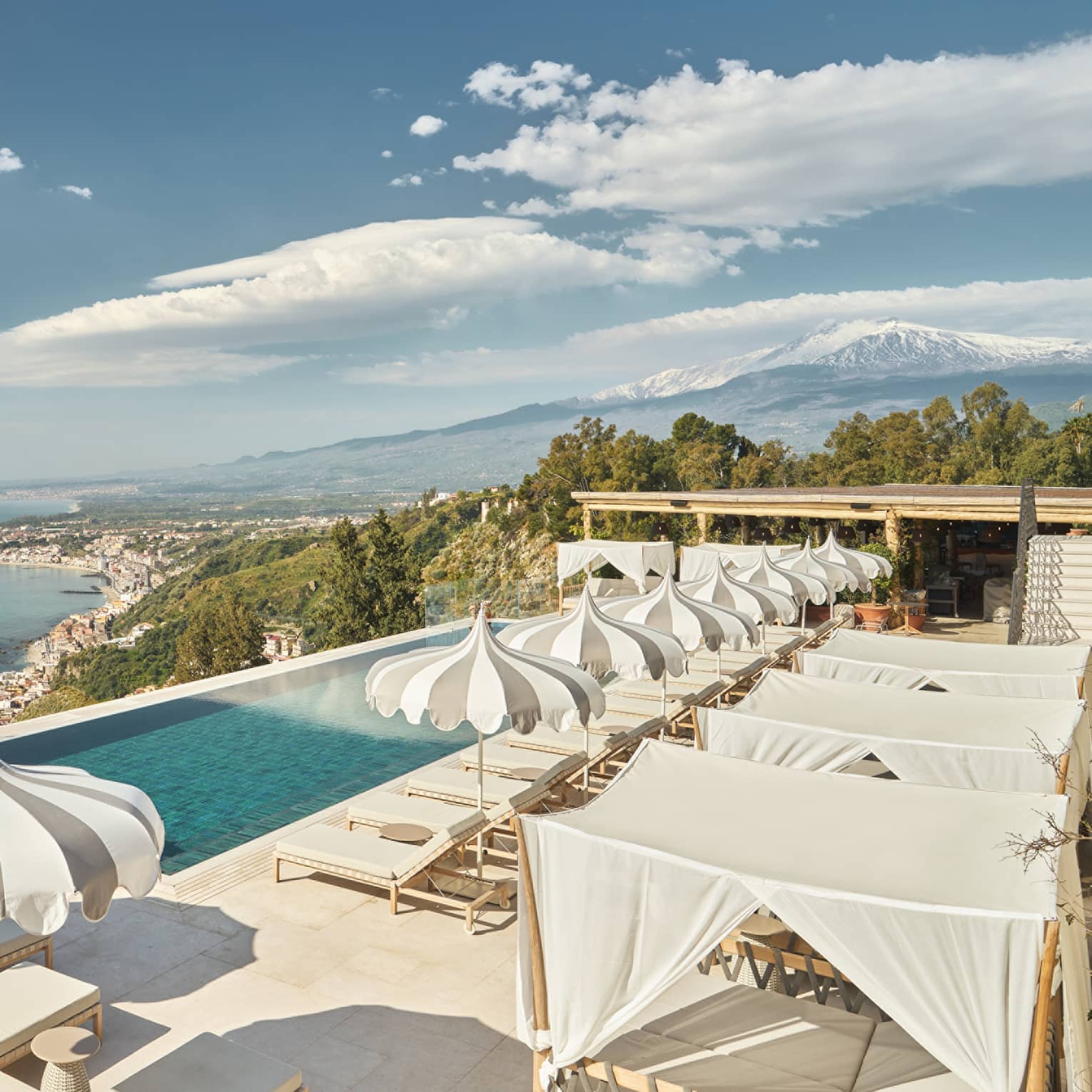 Infinity pool overlooking the sea shore surrounded by grey and white pinstripe pool umbrellas and loungers.