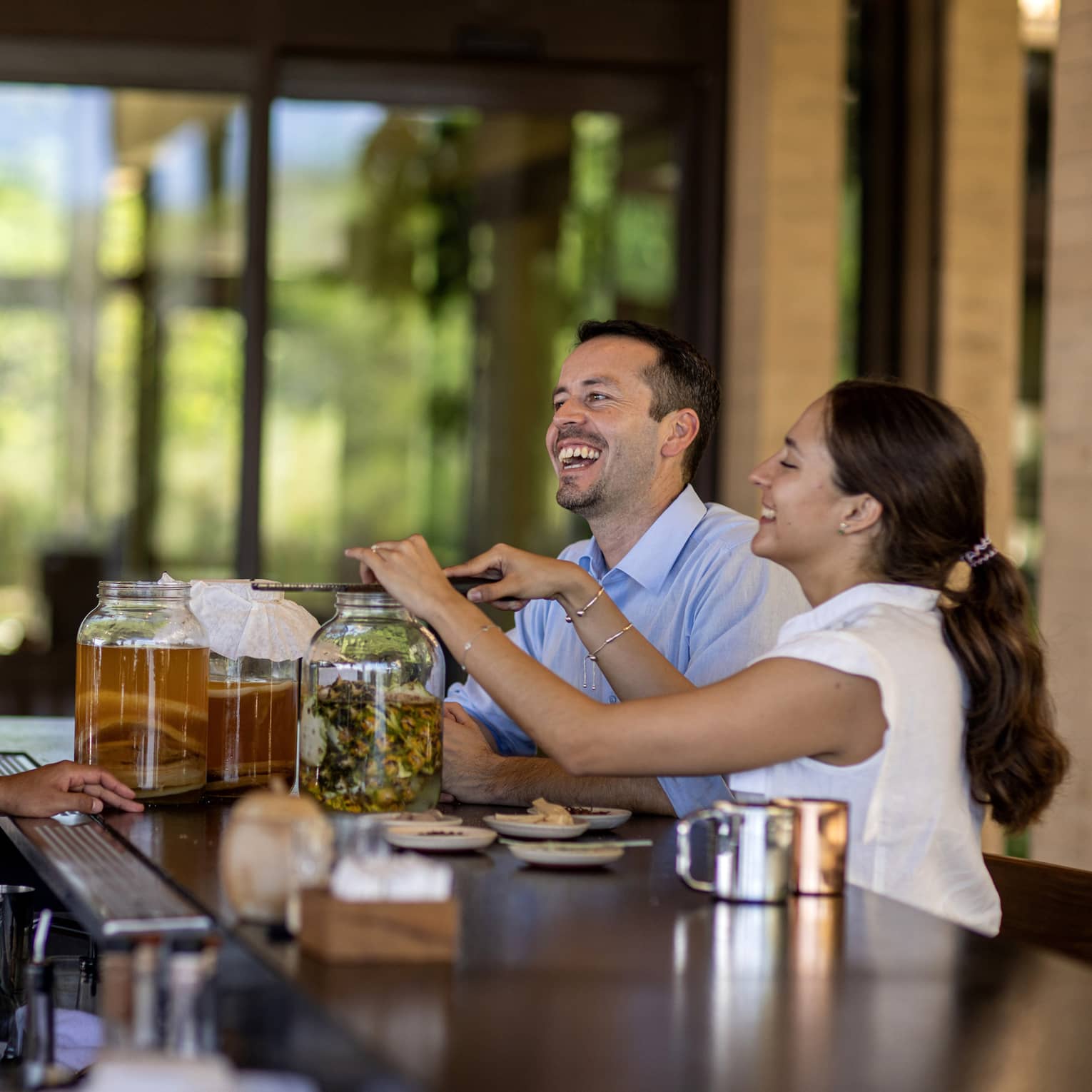 Three guests gather at a bar, one grates an ingredient into a large fermentation jar aside two other jars of amber liquid.