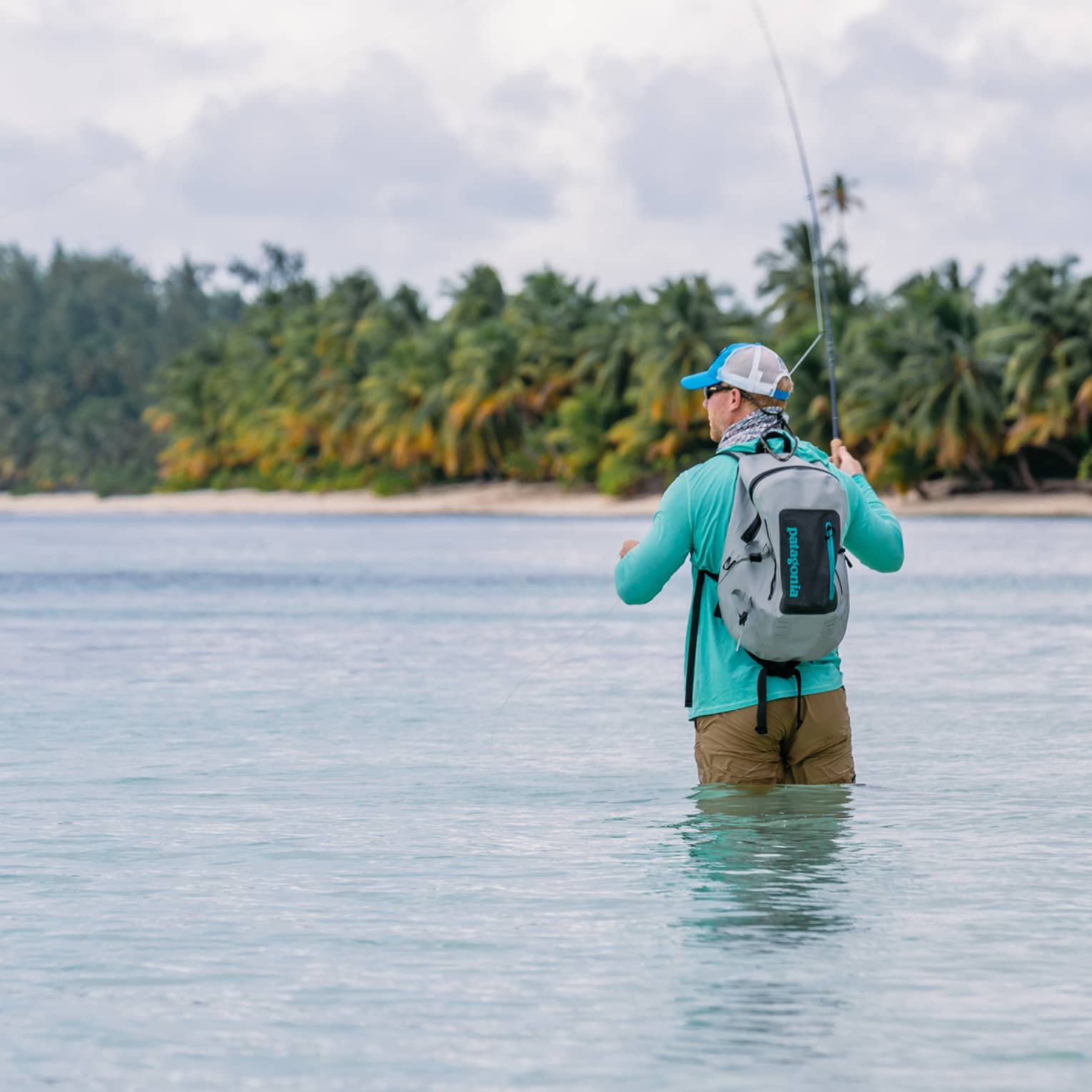 Rear view of fisherman, legs submerged, holding a curved, taut fishing line, distant beach and swaying palms beyond.