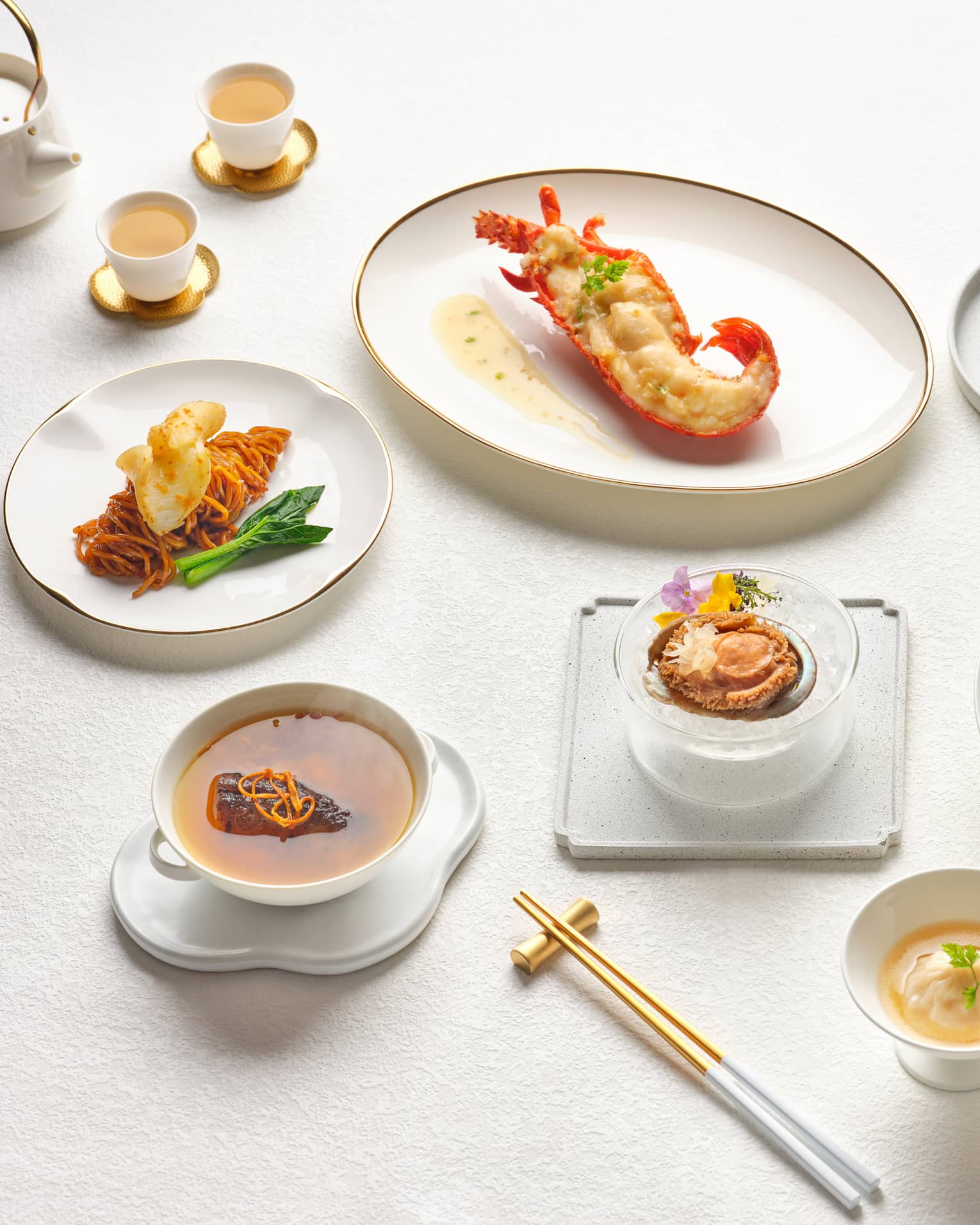 A beautifully arranged gourmet Chinese meal featuring lobster, braised meat, dumplings and dessert, presented on elegant white tableware with teacups and gold chopsticks.