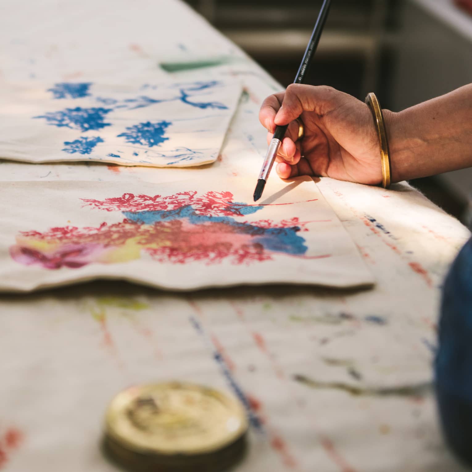 Close-up of a gold-bangled hand wielding a long brush to paint an intricate pattern on a cloth tote bag.