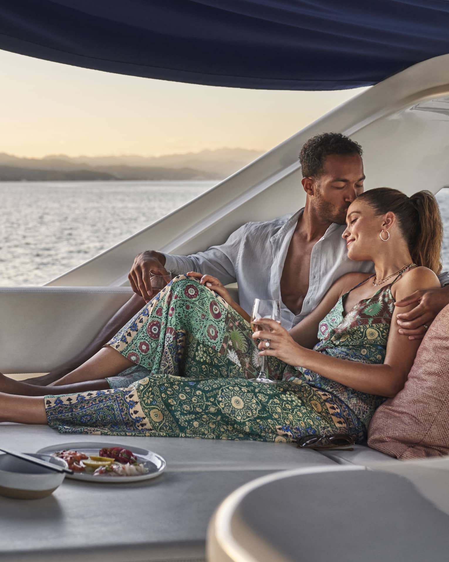 At sunset, a couple lounges in a luxury yacht set with a wine cooler, a plate of lemon slices and a dish of vibrant seafood.