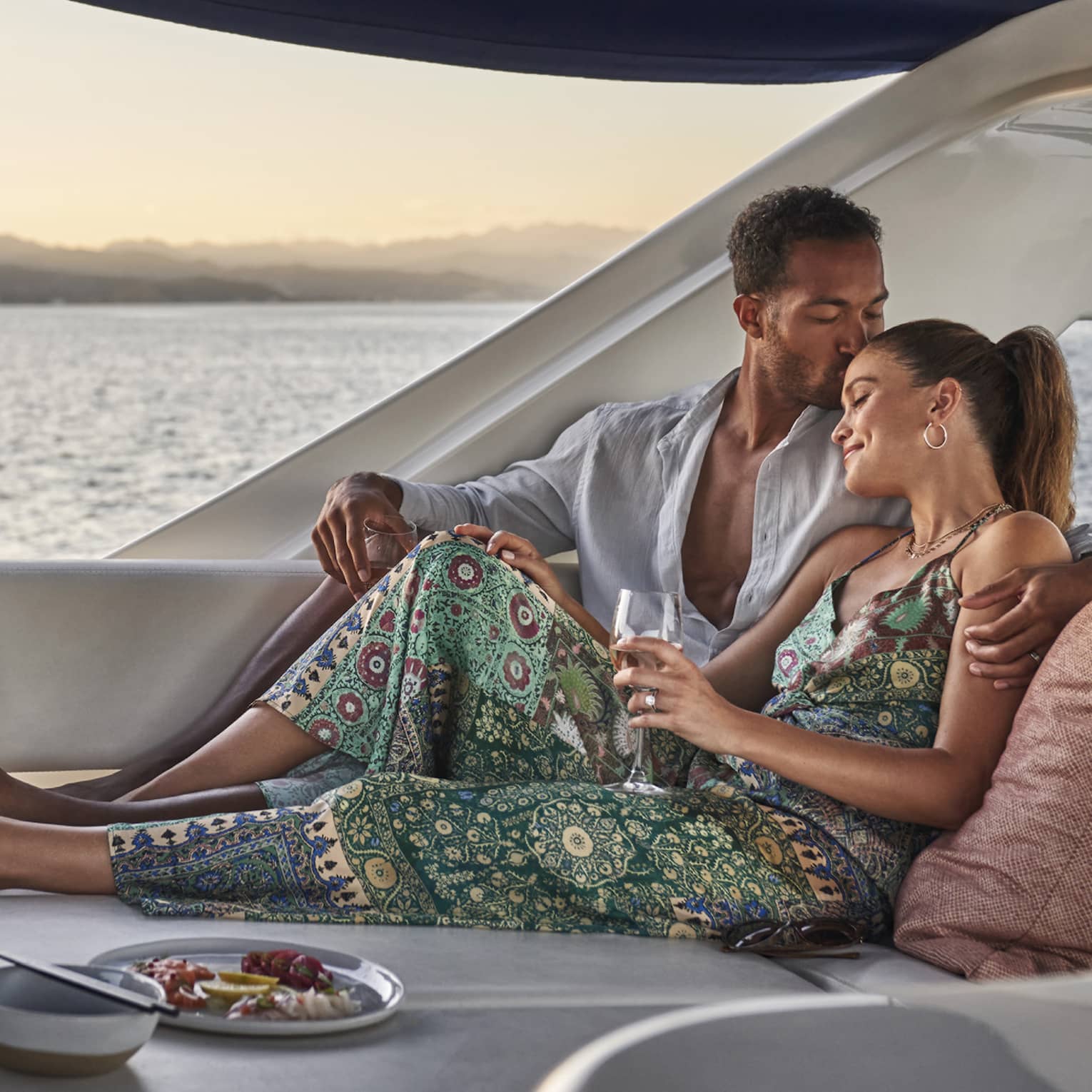 At sunset, a couple lounges in a luxury yacht set with a wine cooler, a plate of lemon slices and a dish of vibrant seafood.