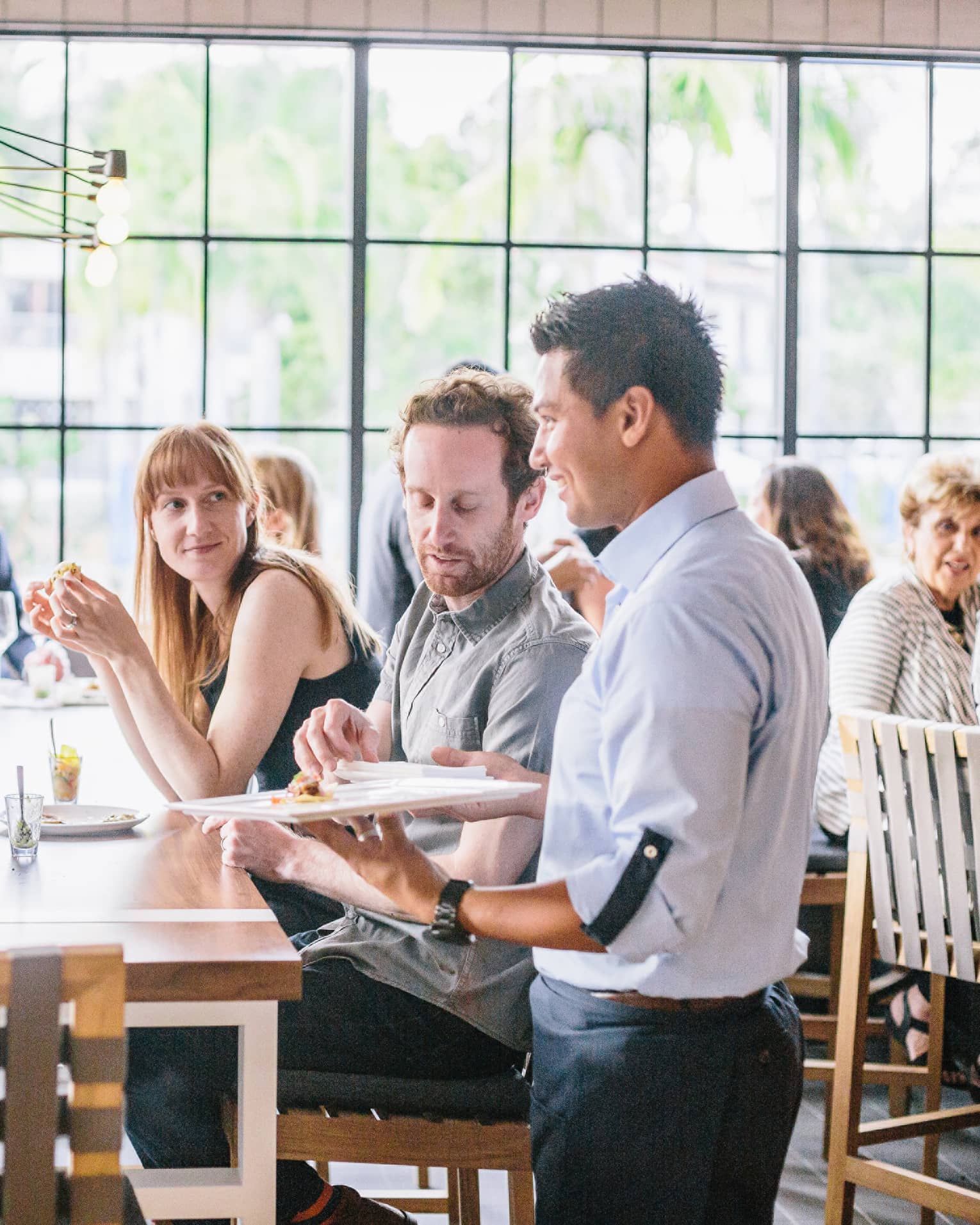 Crowds gathered around long dining table in sunny Seasons Restaurant as server stands with tray of appetizers
