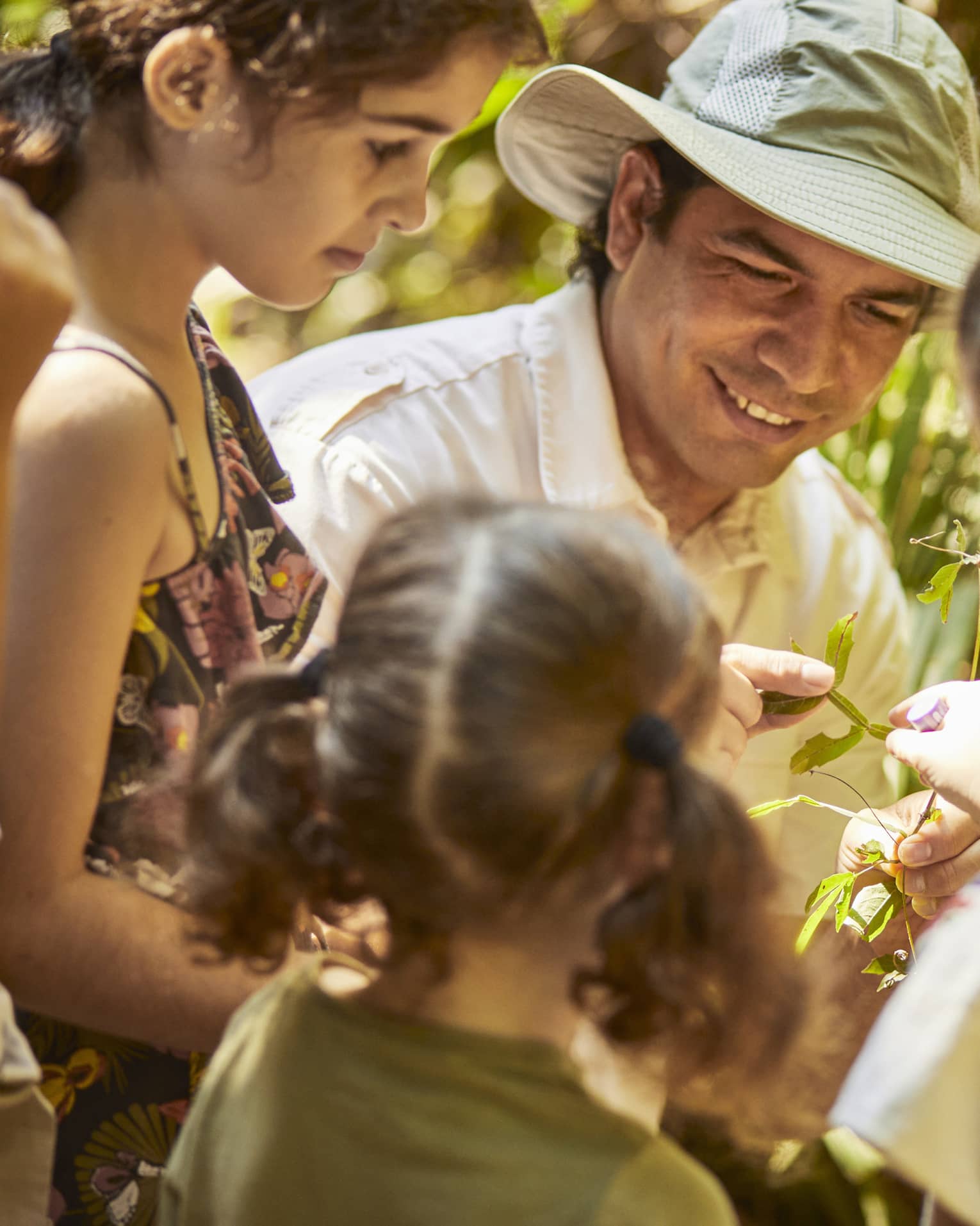 Three children gather around a guide wearing a sun hat as he shows them a plant. One child reaches to touch the leaves.