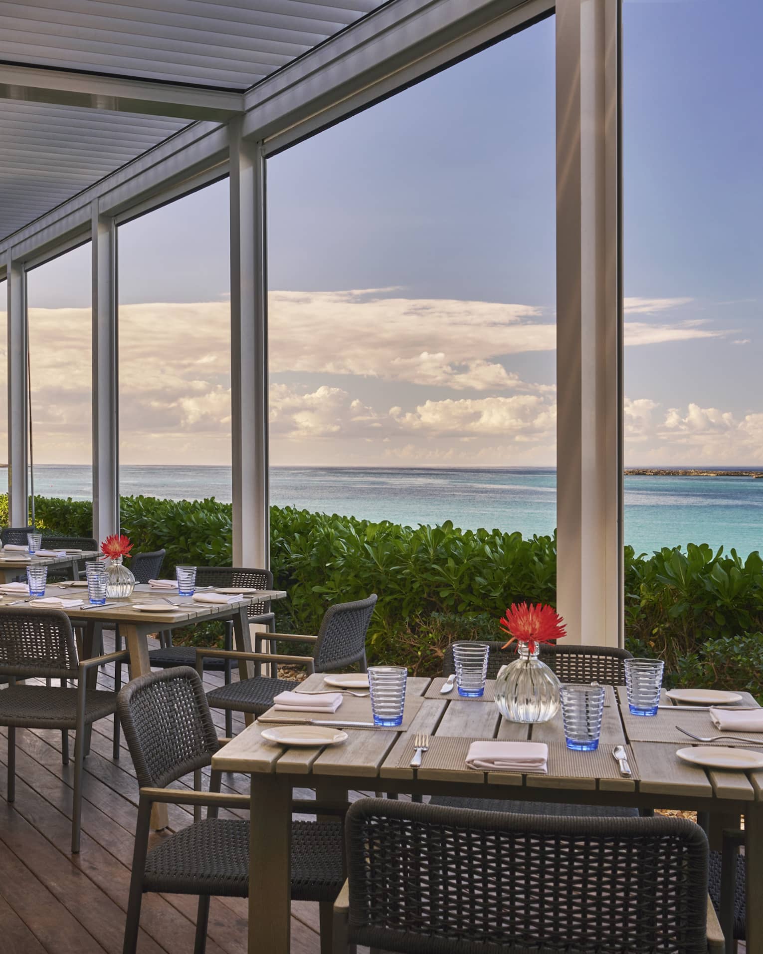 An outdoor eating area with square wood tables next to plants and the ocean.