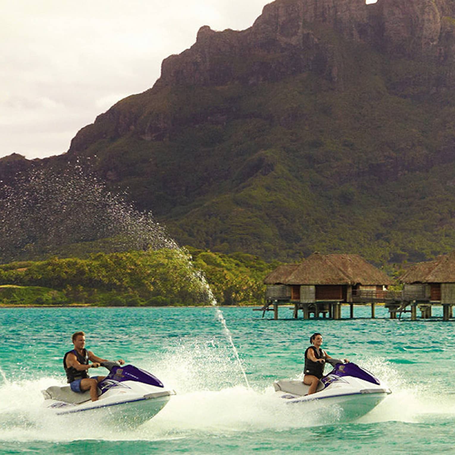 Two jet skis ride along lagoon past row of overwater bungalows, Bora Bora mountain