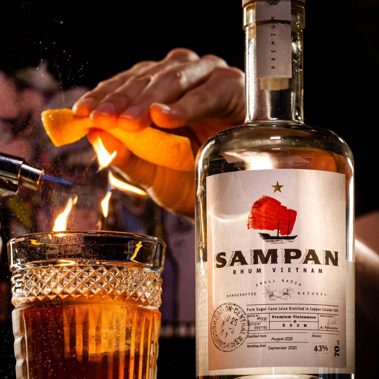 Person ignites an orange peel with a torch over a glass, bottle of Sampan rhum next to it