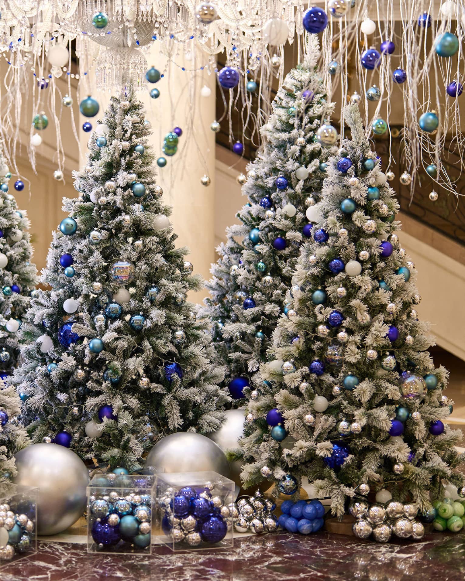 A lobby is decorated with hanging white garland and five frosted Christmas trees adorned with blue and silver ornaments.