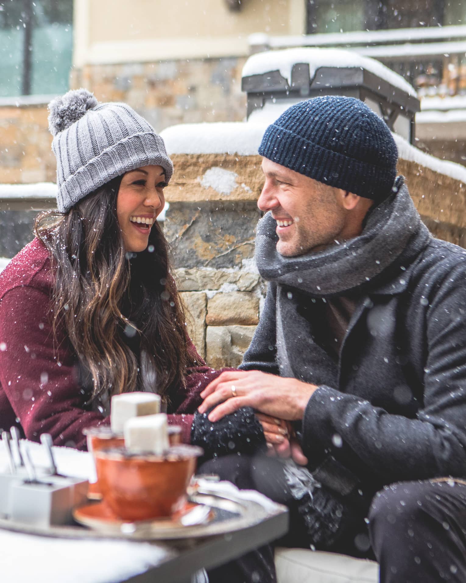 Smiling couple sitting by a firepit in a snowy outdoor setting, bundled in winter coats and hats. Snowflakes fall around them as they enjoy a cosy moment. 