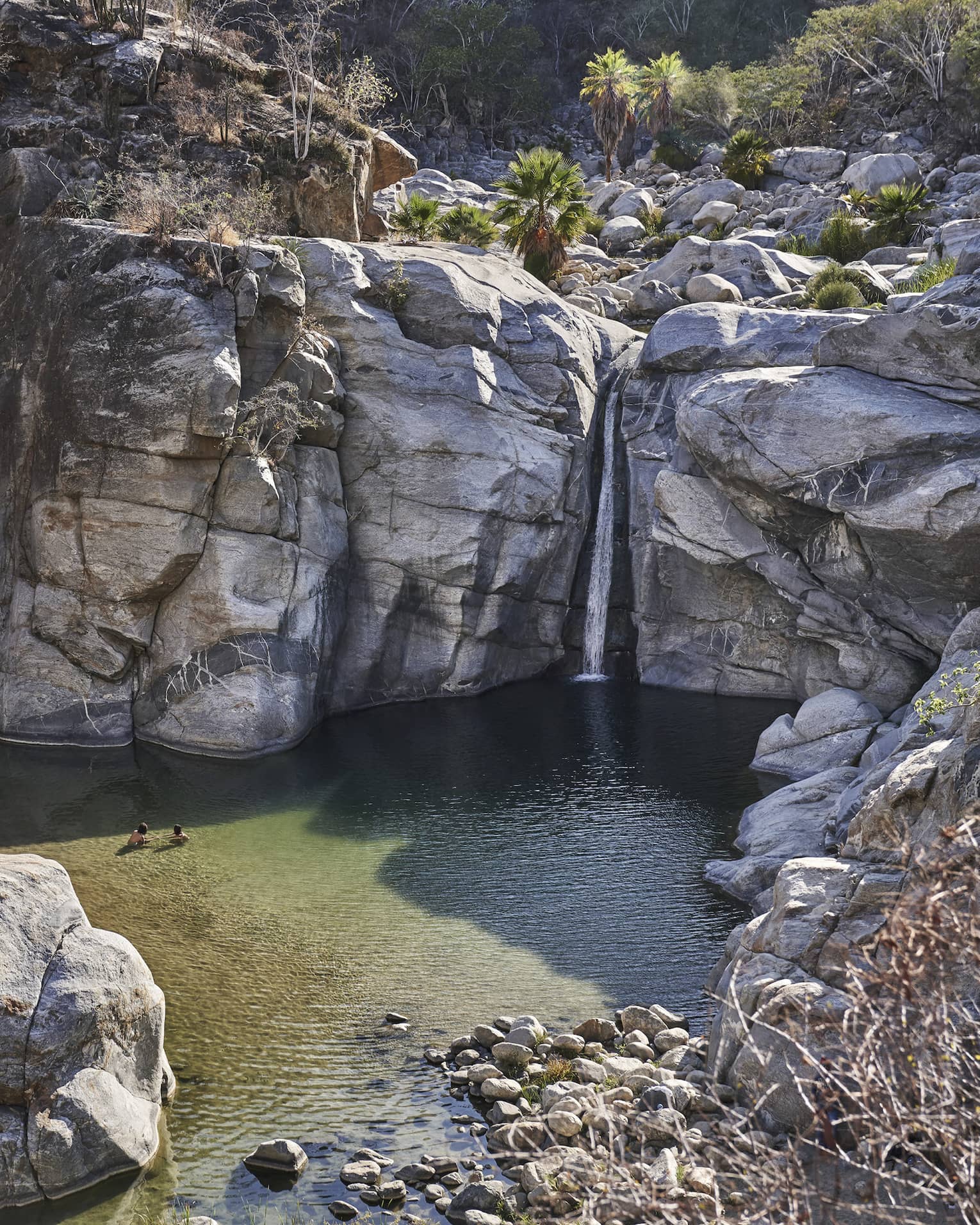 Two people wade in a small ocean lagoon surrounded by large cliffs, creating a serene and picturesque scene with clear water and rugged natural beauty.