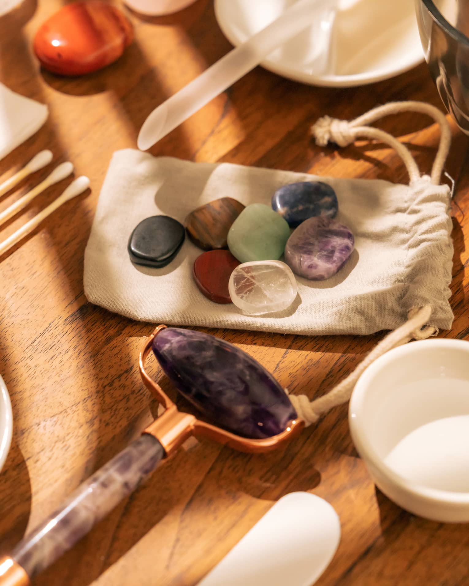 Face roller, gemstones, cotton swabs and a variety of small white bowls and other instruments sit on a sun-lit wooden table