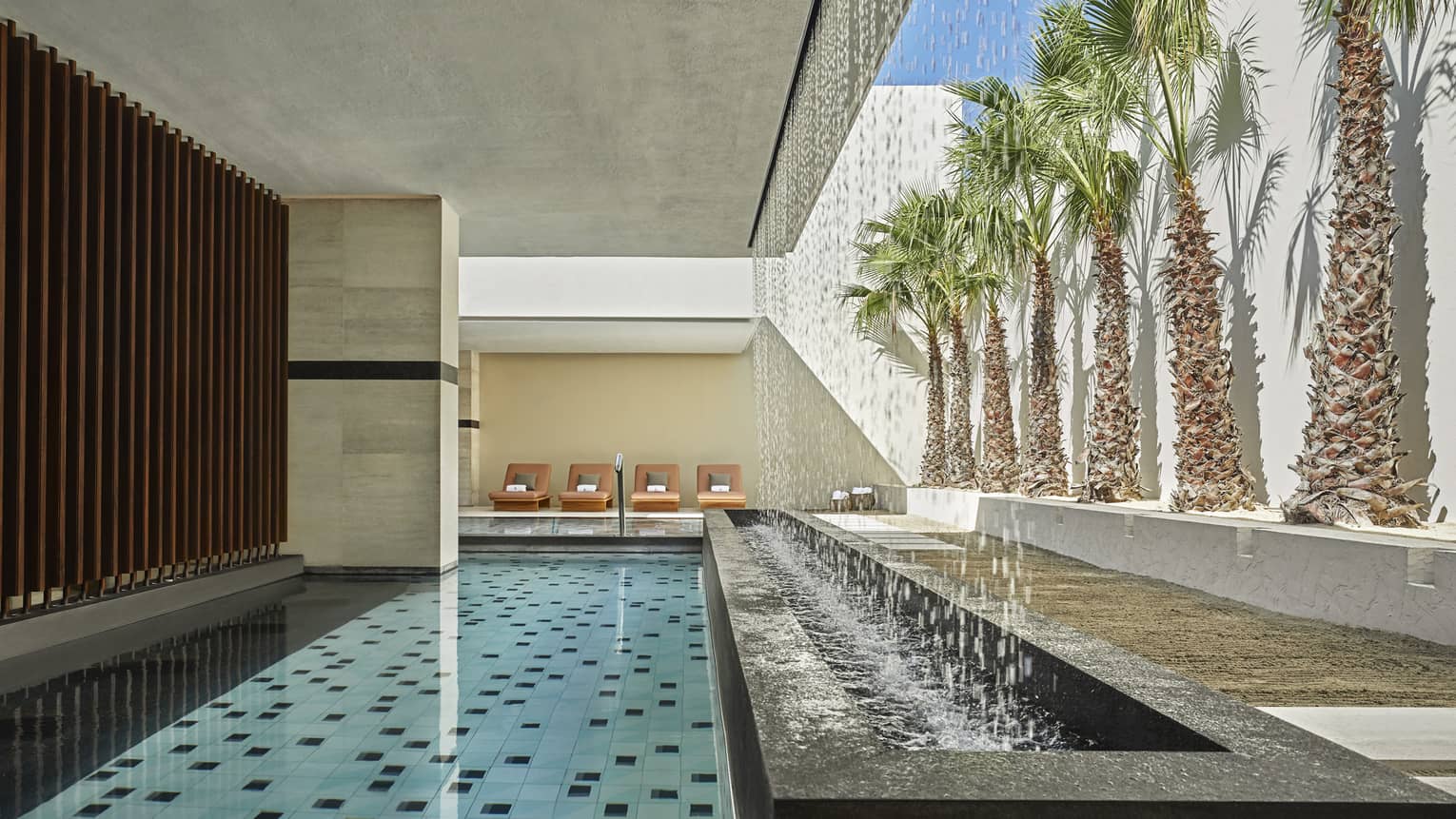 An outdoor pool is covered with a white stucco overhang and lined with lounge chairs on one side, palm trees on the other 
