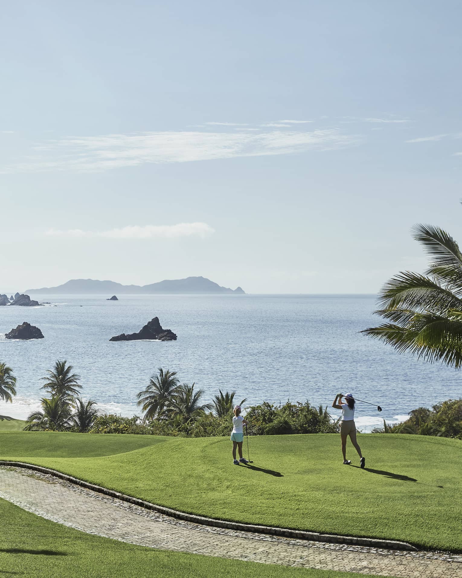 A guest swings from the tees on a hilly fairway bordered by palm trees and blue water with rocky outcrops as a child watches.