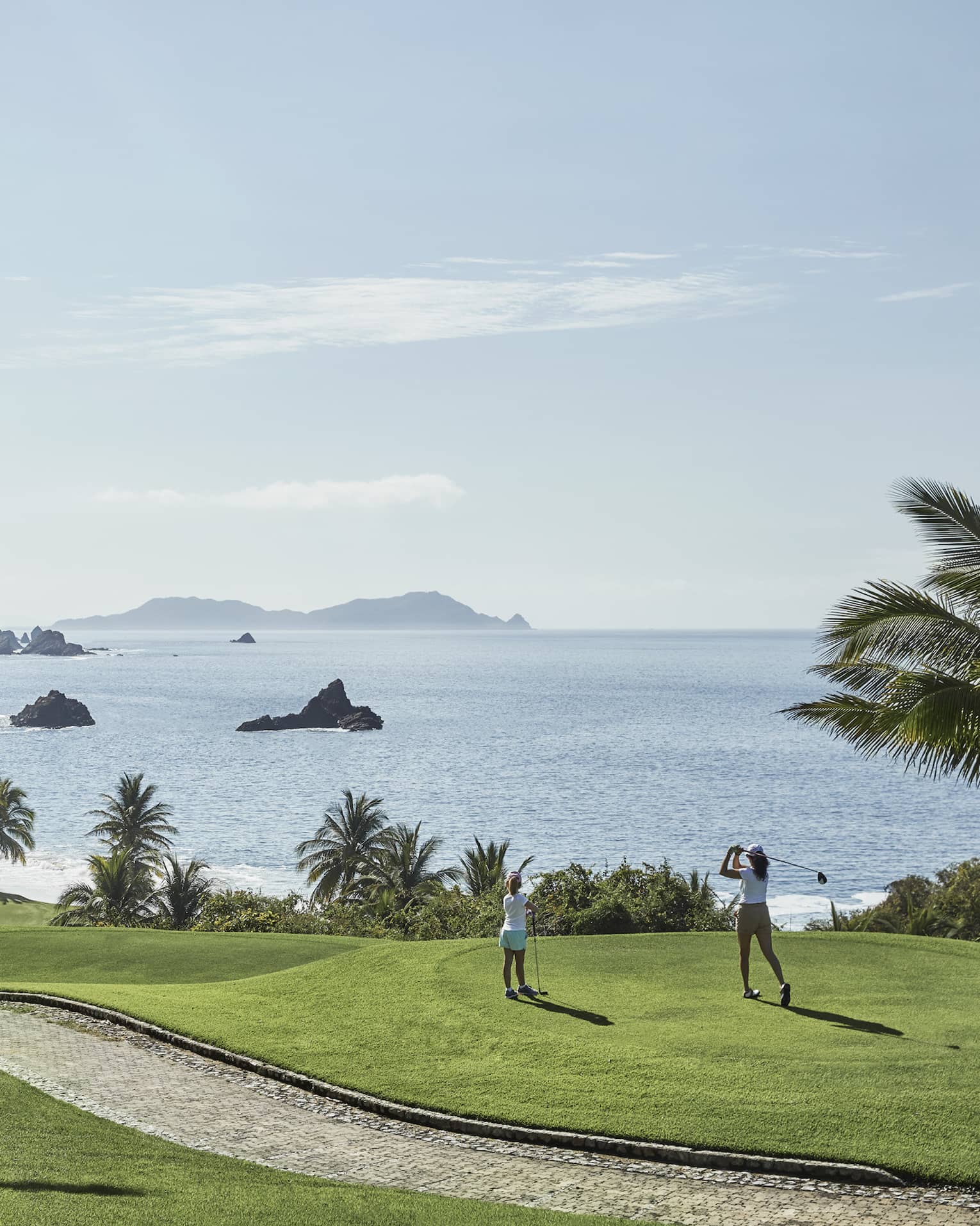 A guest swings from the tees on a hilly fairway bordered by palm trees and blue water with rocky outcrops as a child watches.