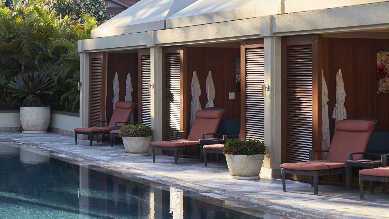 Cabanas line outdoor swimming pool deck under palm trees and sunny sky
