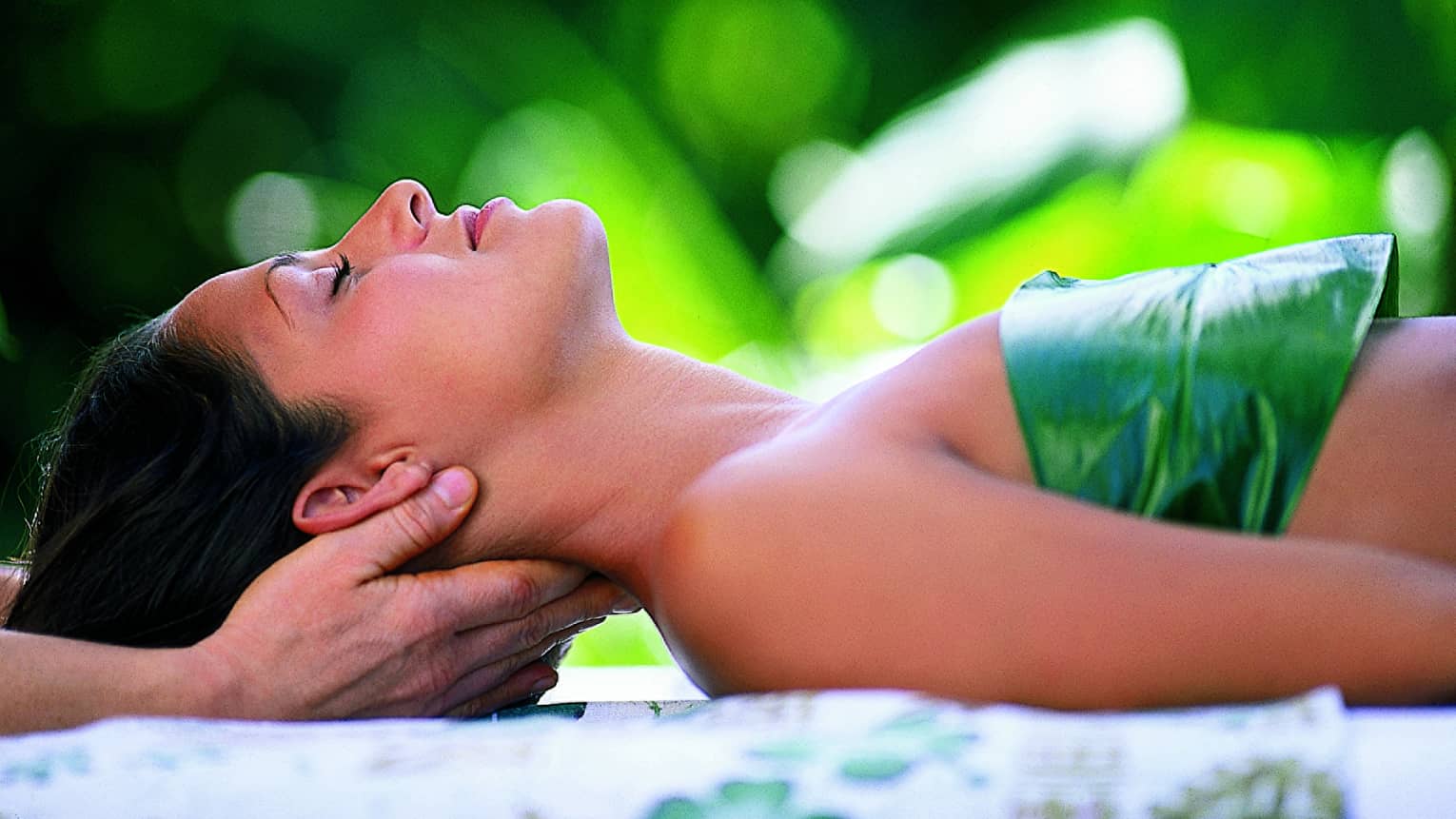 Woman with eyes closed lays on back on table with leaf over chest, hands massage her neck