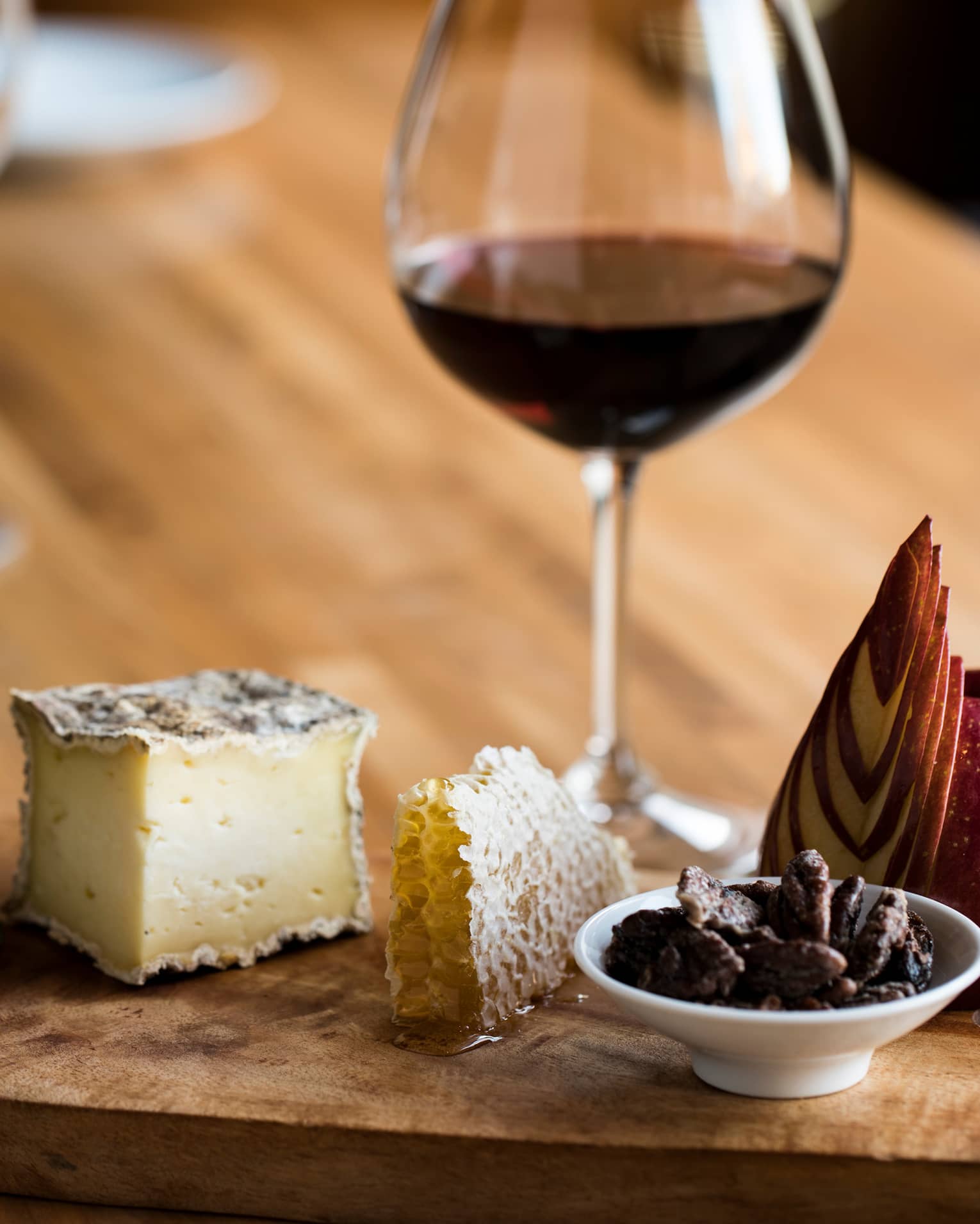 A glass of red wine with a luxurious wooden tray holding cheese, honeycomb, honey, sliced apple, nuts and crackers.
