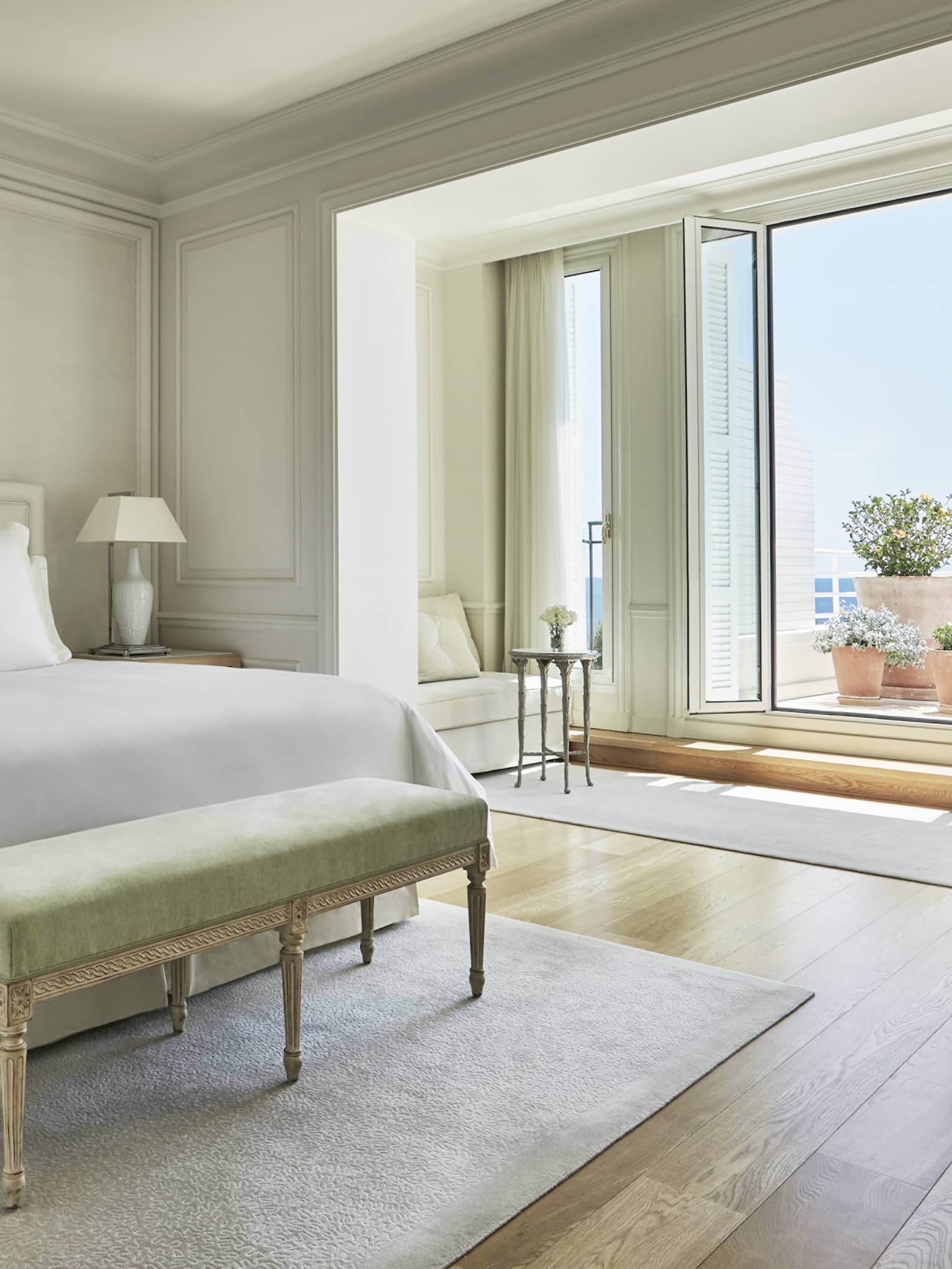 Hotel suite with white bed and green foot bench on neutral rug with terrace and sea views from tall glass terrace doors