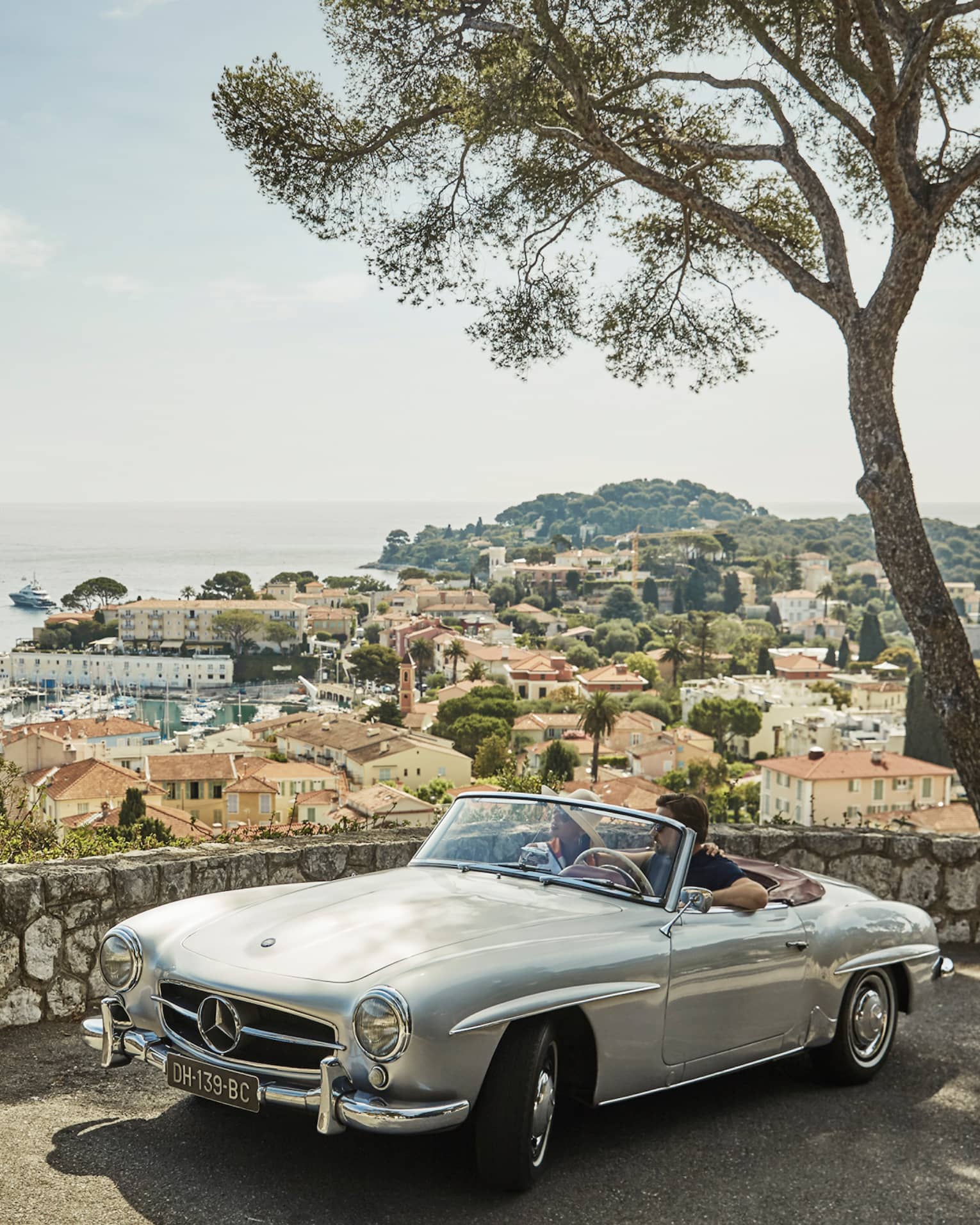 Couple in luxury vintage white car parked under tree by stone wall overlooking red roofs, water