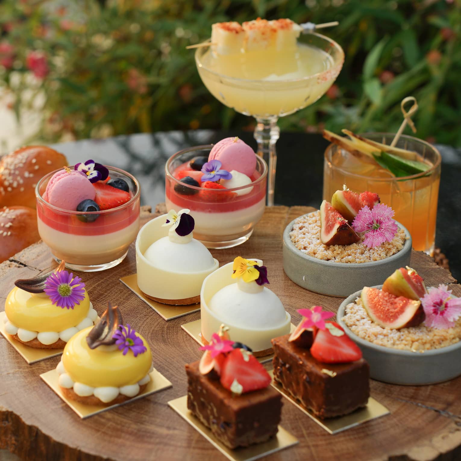 Tray of small decadent desserts in varying shapes and colours topped with fruits and floral accents, served with cocktails.