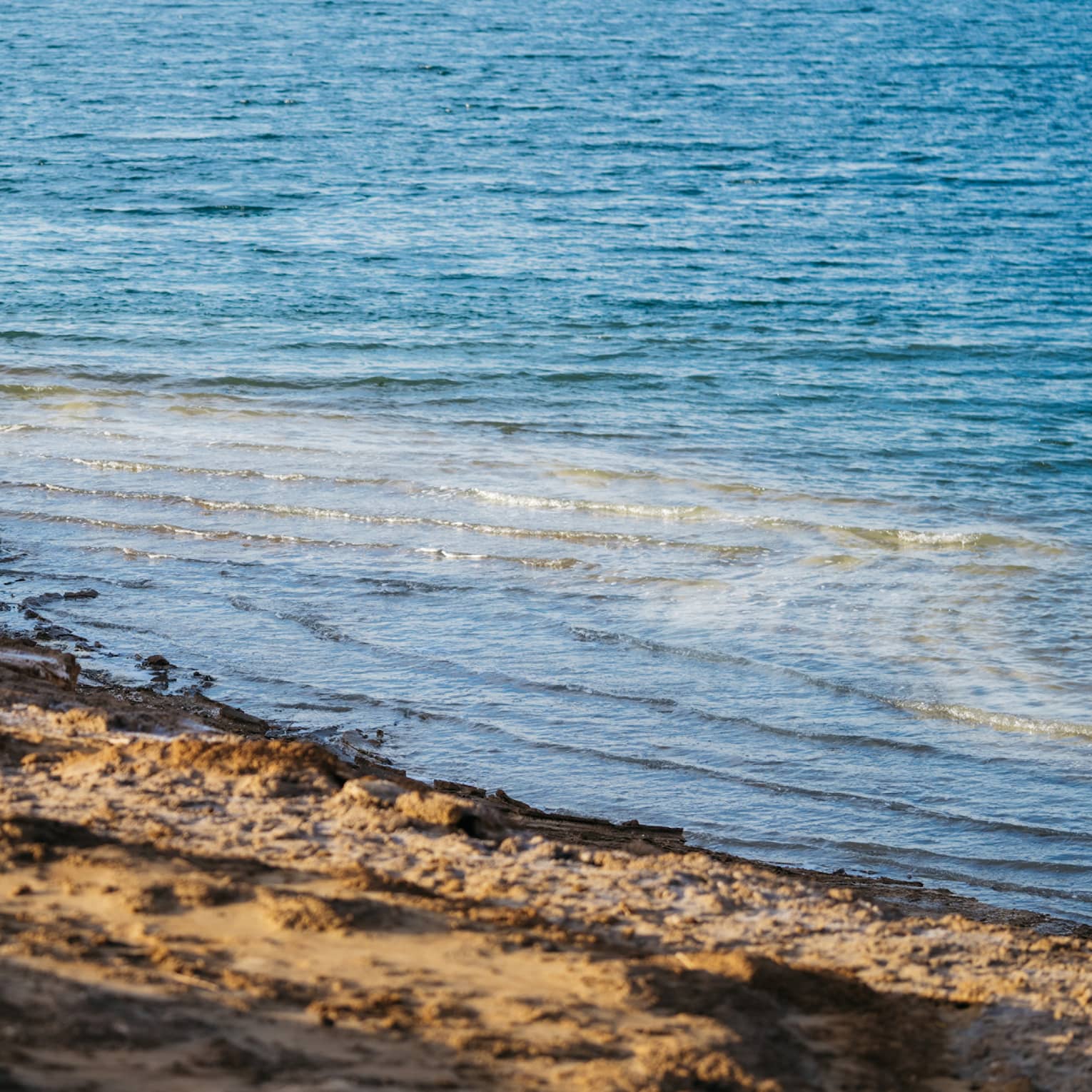 Gentle waves in sparkling azure water undulate toward an empty dark-brown, rough-sand beach.