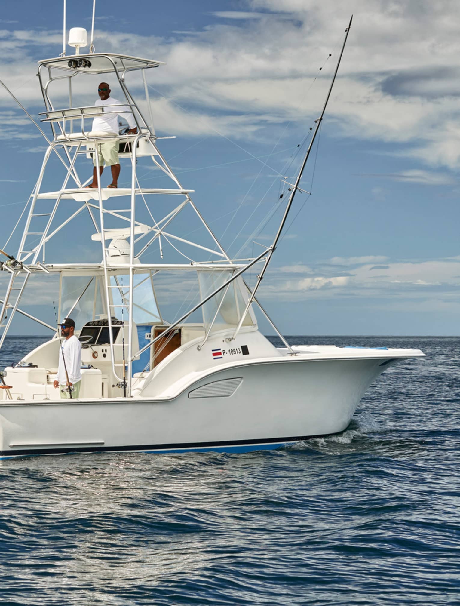 A group of people on a white fishing boat in calm ocean waters, with fishing rods ready. One person is standing on the upper deck, and the sky is partly cloudy.