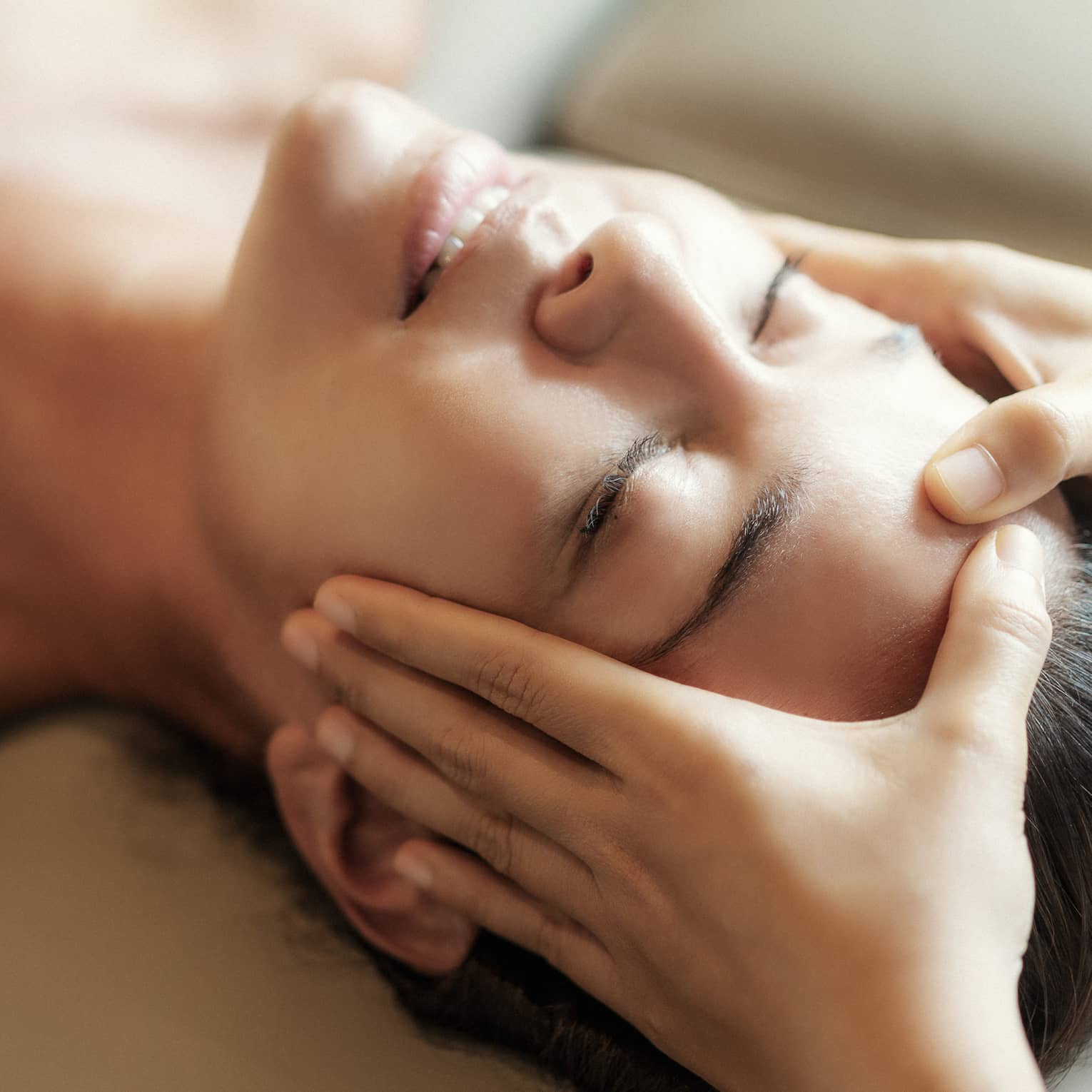 Spa facial, two hands rest on woman's forehead as she closes her eyes, lays under sheet on treatment table