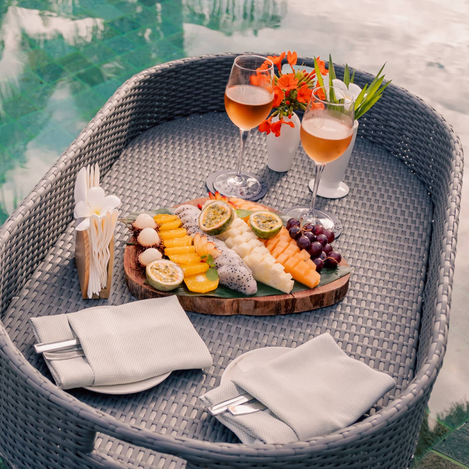 A grey wicker tray floats in a pool carrying a board of fruit arranged in rows, two plates and two glasses of rosé.