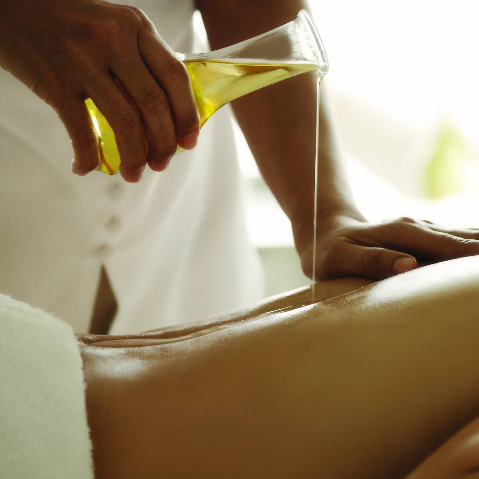 Spa staff pours massage oil over woman's bare back as she lays on treatment bed