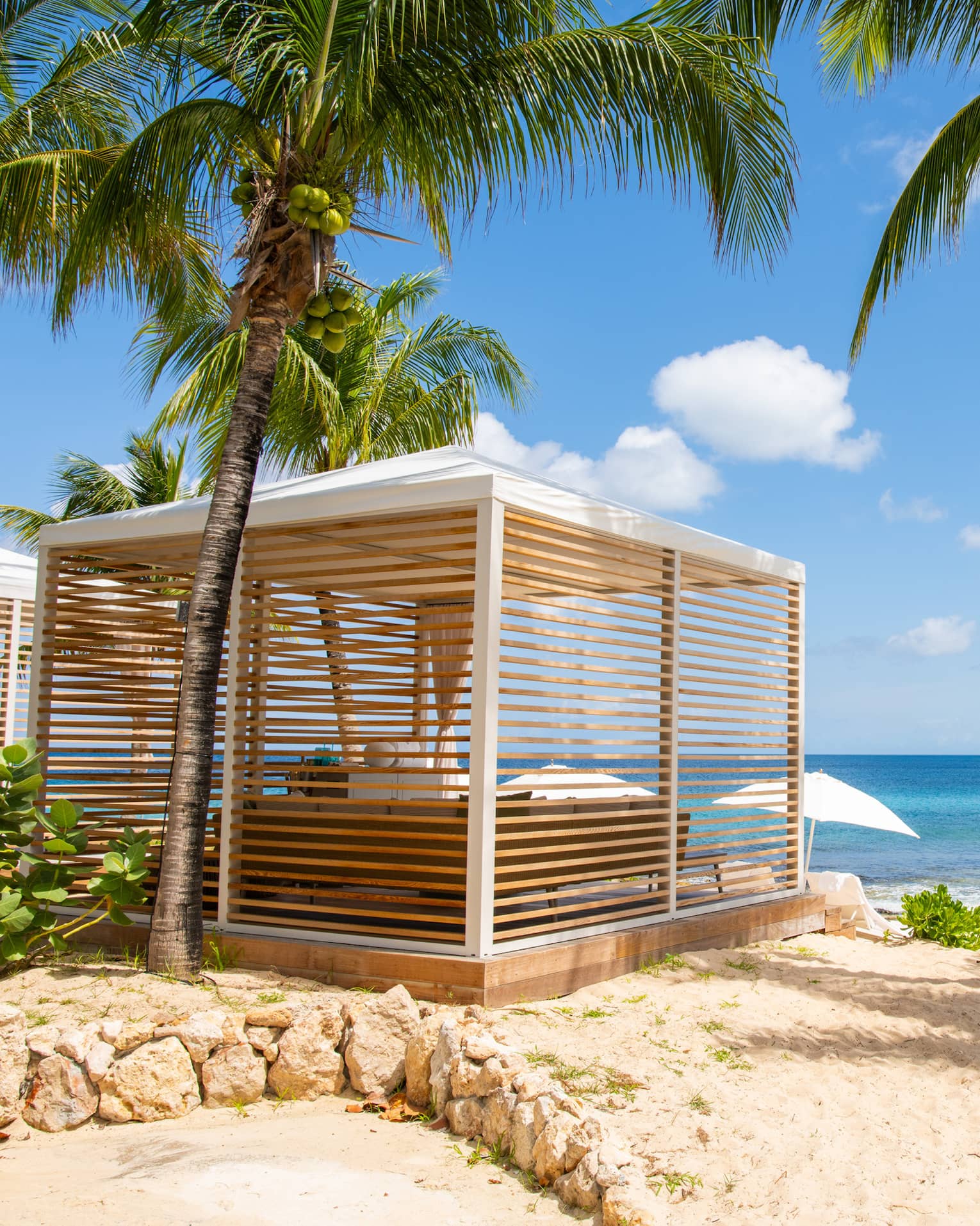 Luxury resort cabana with wooden slats surrounded by palm trees, located on a sandy beach and overlooking the ocean