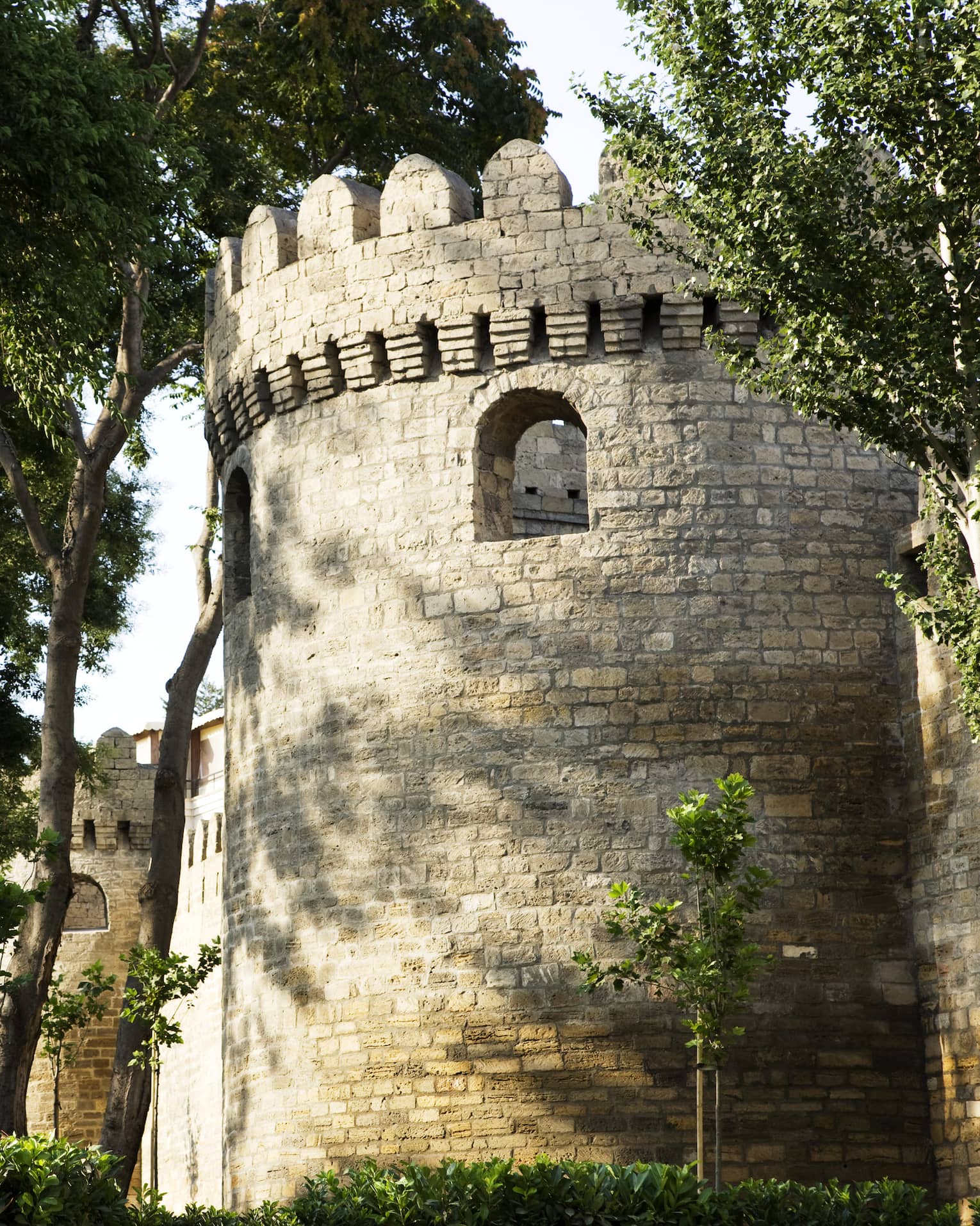 A cylindrical structure marked by loophole openings and grey stone exterior: part of a crenelated tower surrounded by trees.
