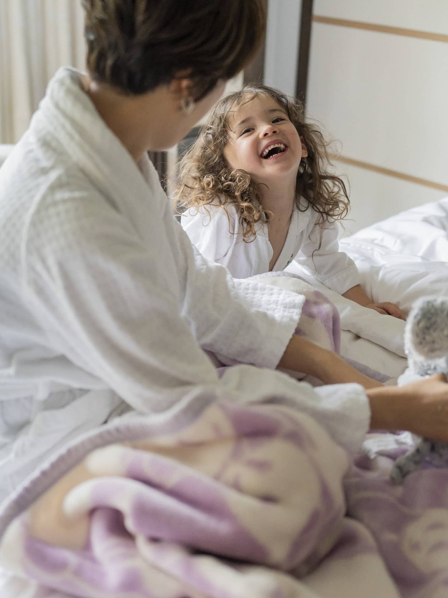 Mother and daughter in matching white bathrobes share a laugh in bed