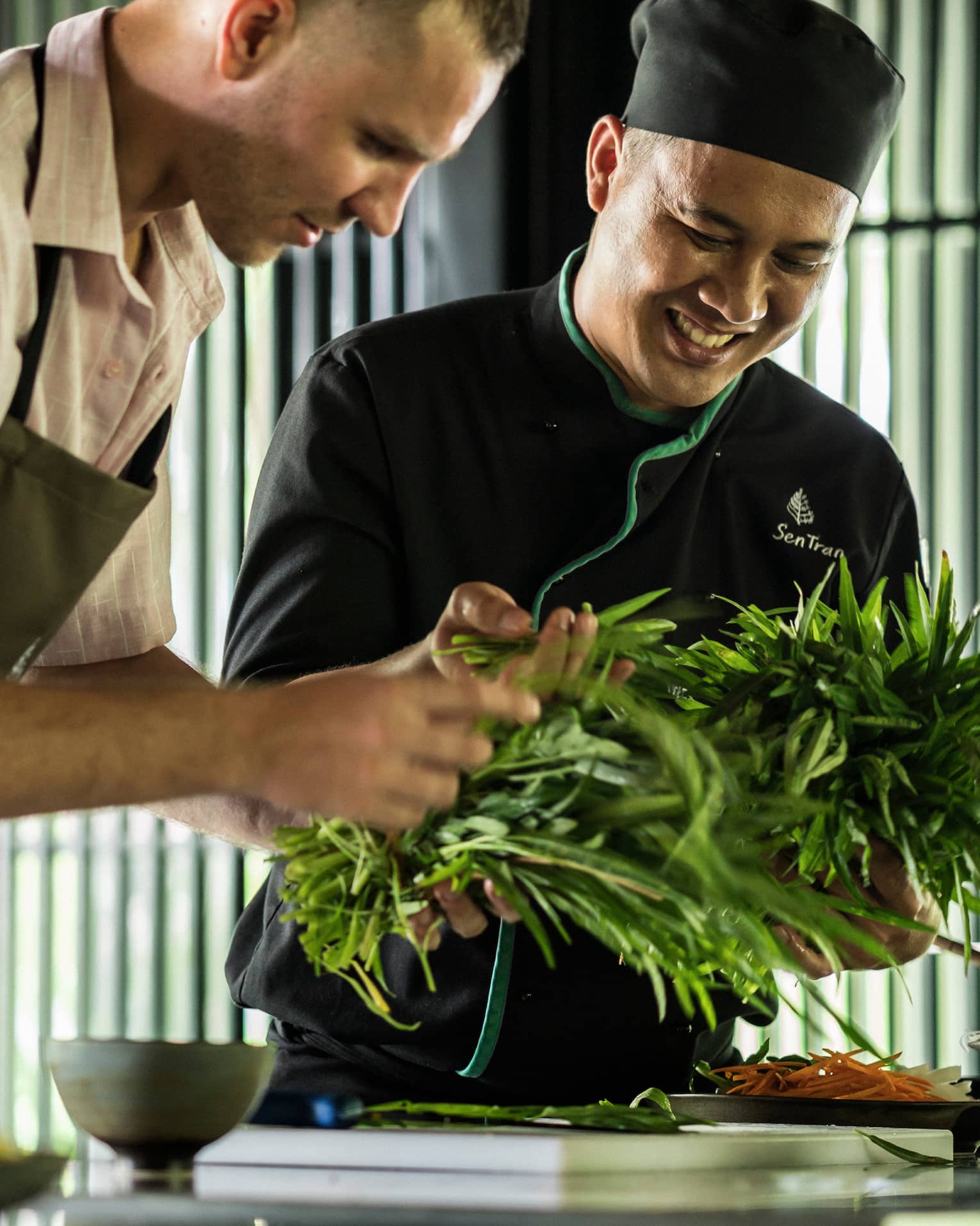 Smiling Chef Tran Van Sen holds greens, shows them to man in apron